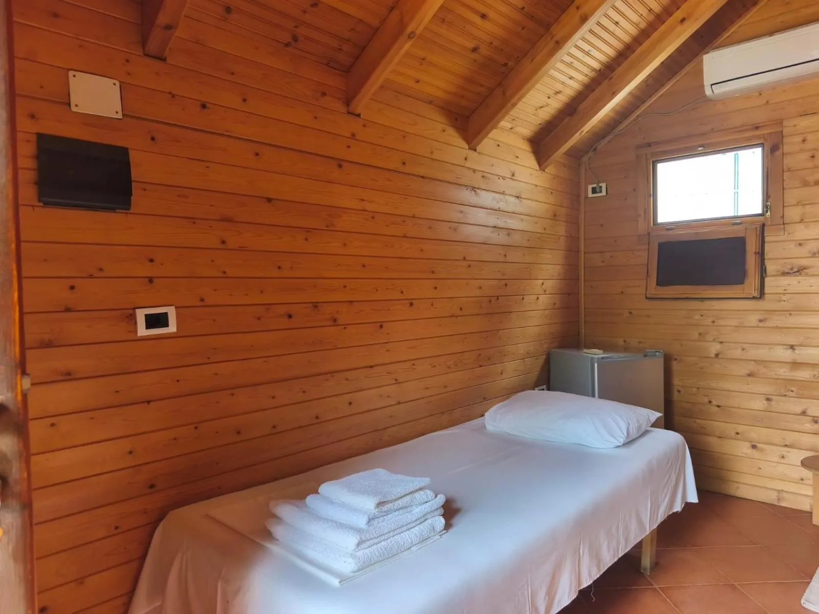 A single bed with folded towels under a small window and an air-conditioning unit in a pine-boarded room