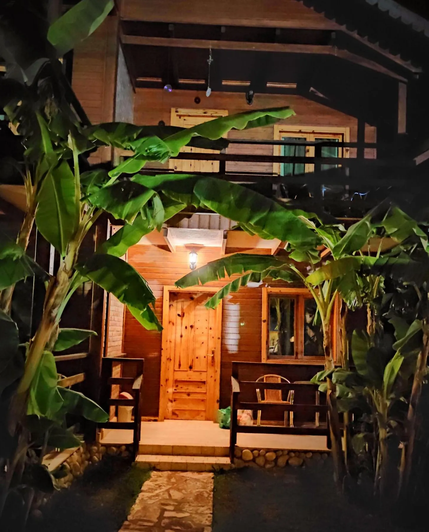 A lit cabin door seen through banana leaves at night, two chairs on the porch