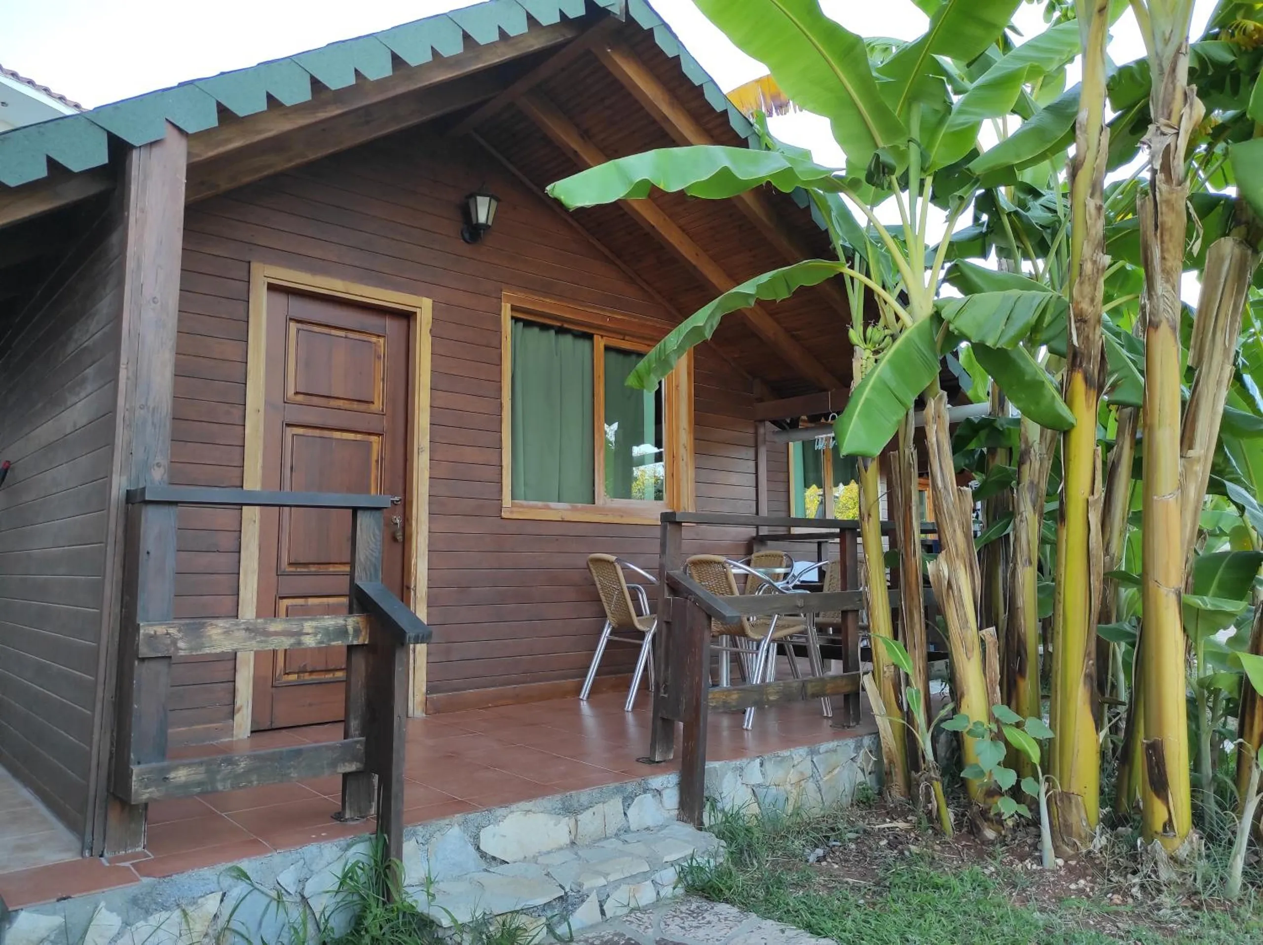 A single-storey timber chalet by day with a pine door, a covered porch with chairs on a stone plinth, and banana stems taller than the roof