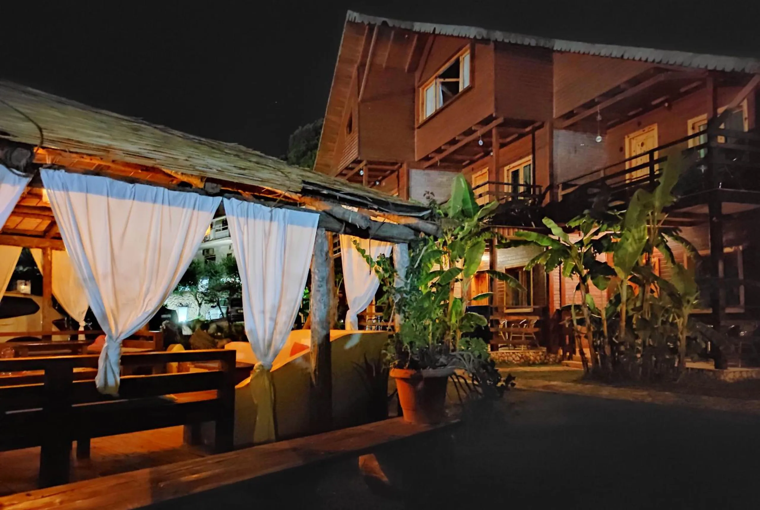 White curtains tied back on the posts of the thatched pavilion at night, the two-storey timber chalet behind it among banana plants