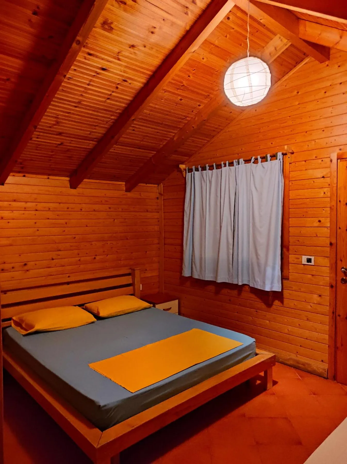 A double bed under the sloping pine ceiling, a curtained window above it