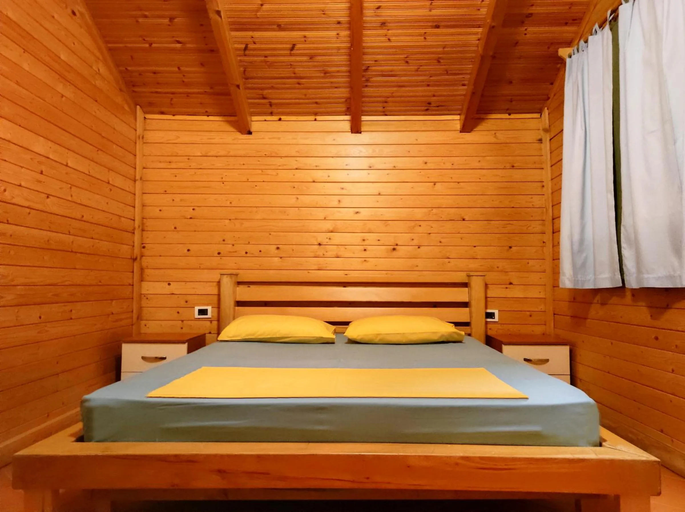 A double bed with a pine headboard in a room boarded in honey pine, a curtained window on the right