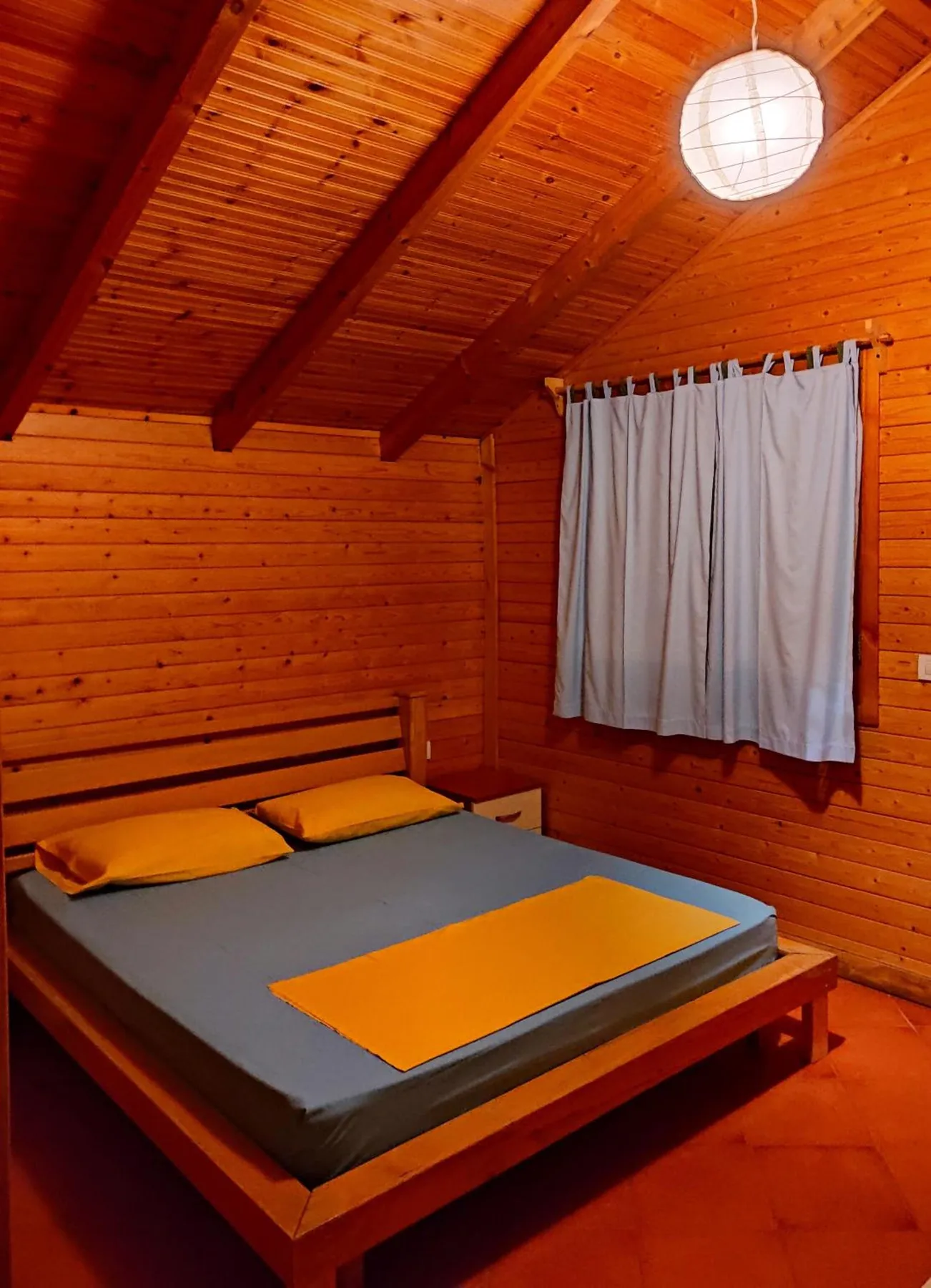 A double bed with grey covers and yellow throws under exposed rafters in a pine-boarded room, a white curtain at the window