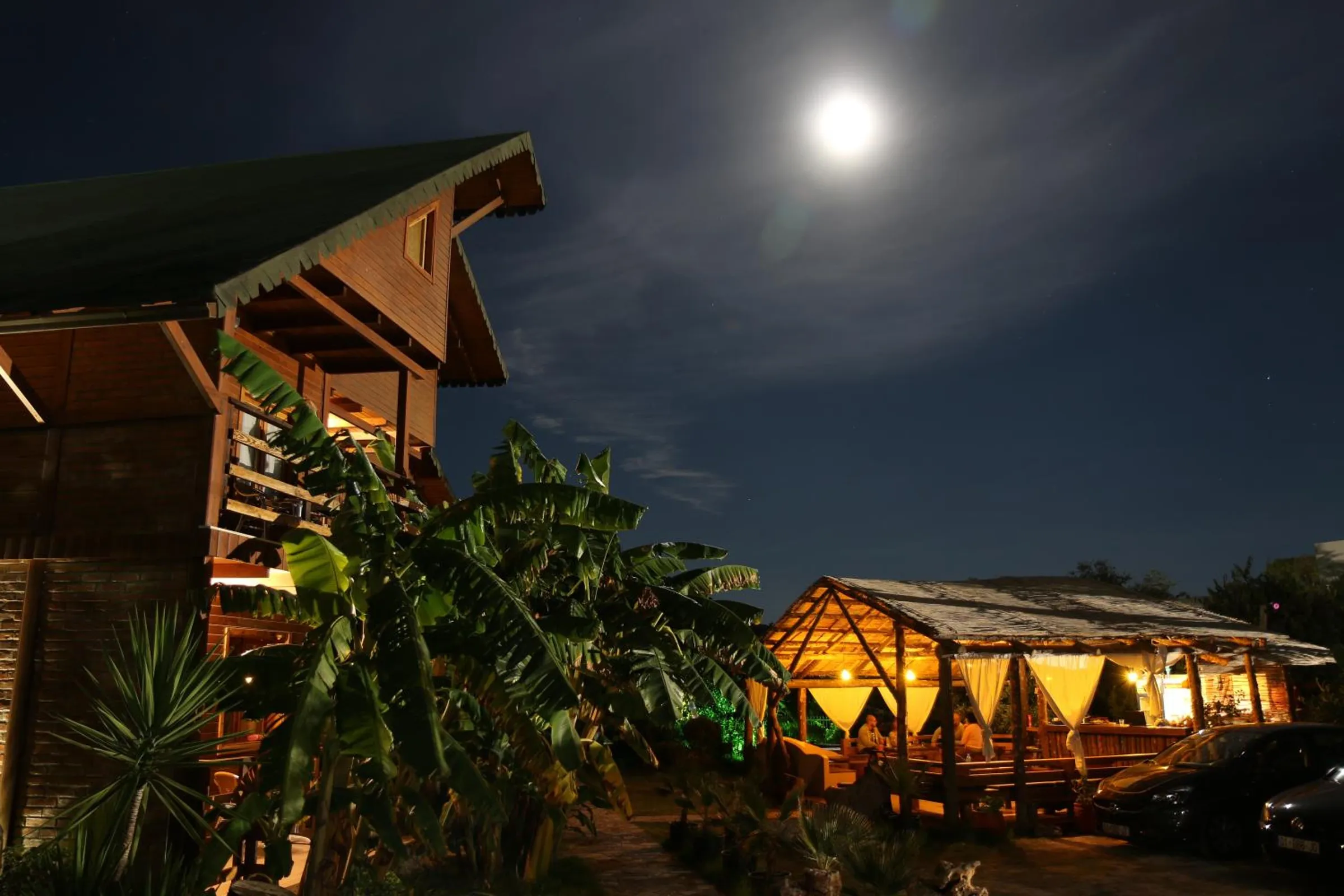 The moon over the garden: a lit chalet balcony on the left, banana leaves, and the pavilion glowing under its thatch on the right