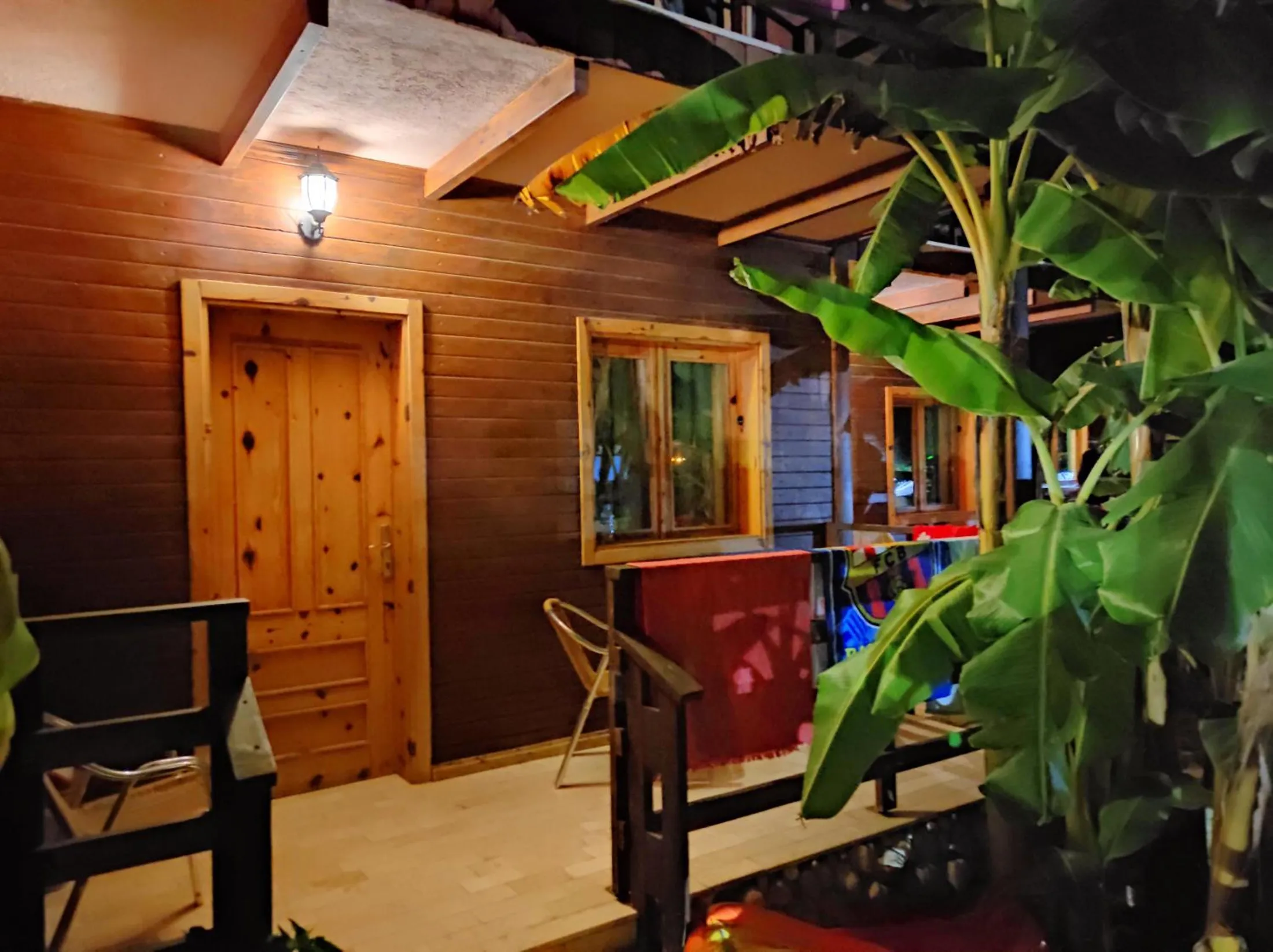 A pine door under a porch lamp at night, a timber bench and a banana leaf in the foreground