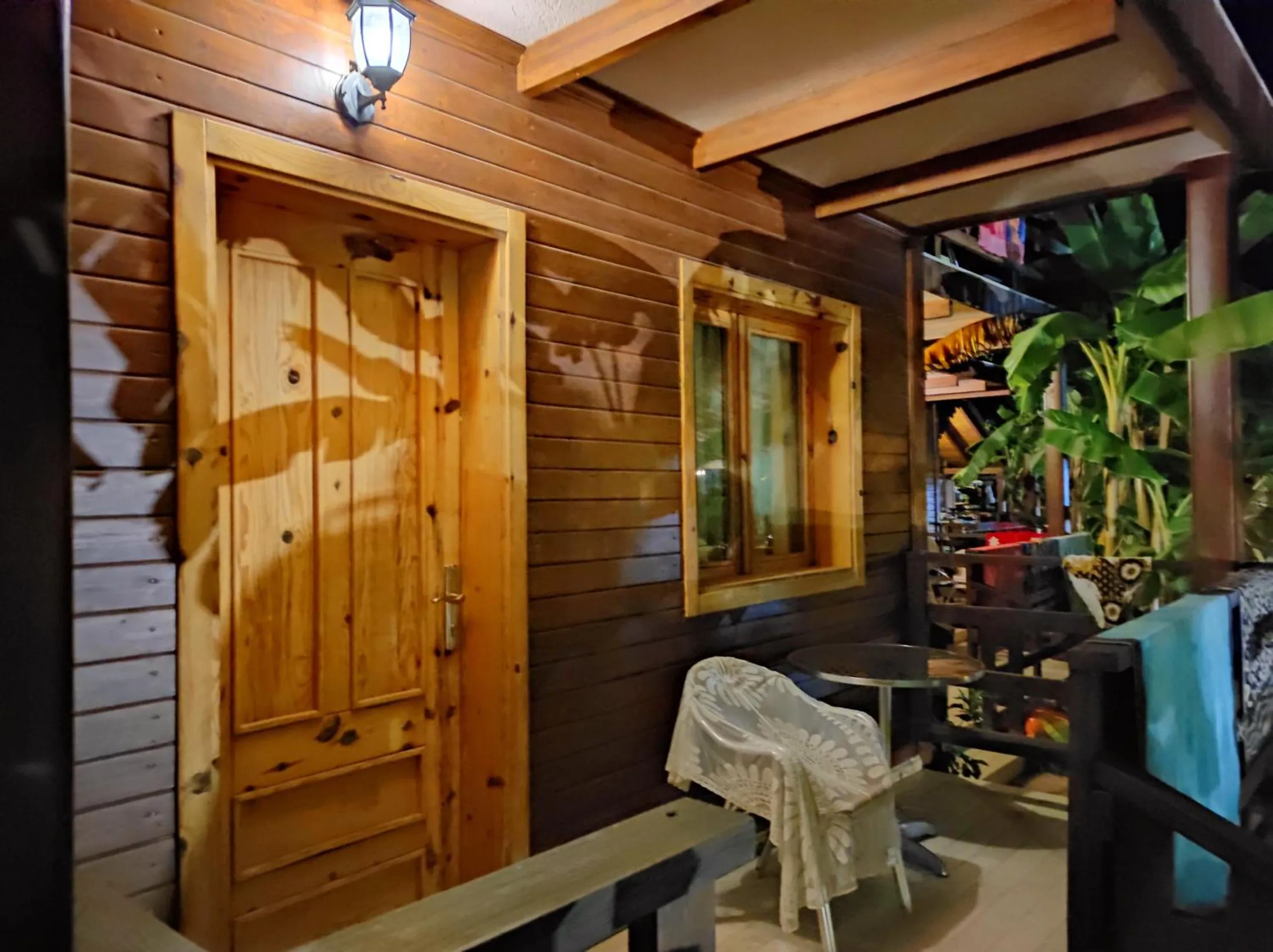 A pine-panelled door and window on a lamplit porch, a cushioned chair and a timber rail in front