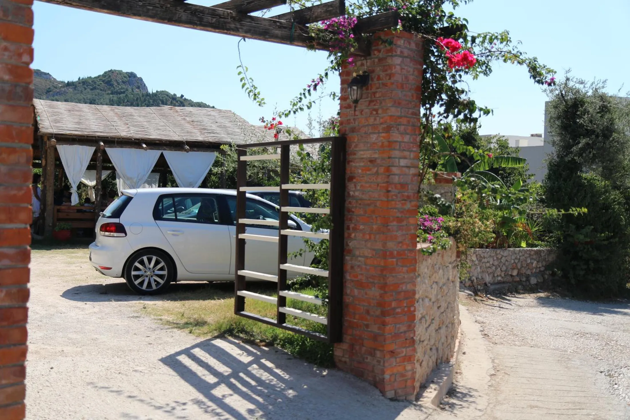 A brick gatepost with bougainvillea and a white gate open onto a gravel yard, a parked car and the curtained pavilion behind