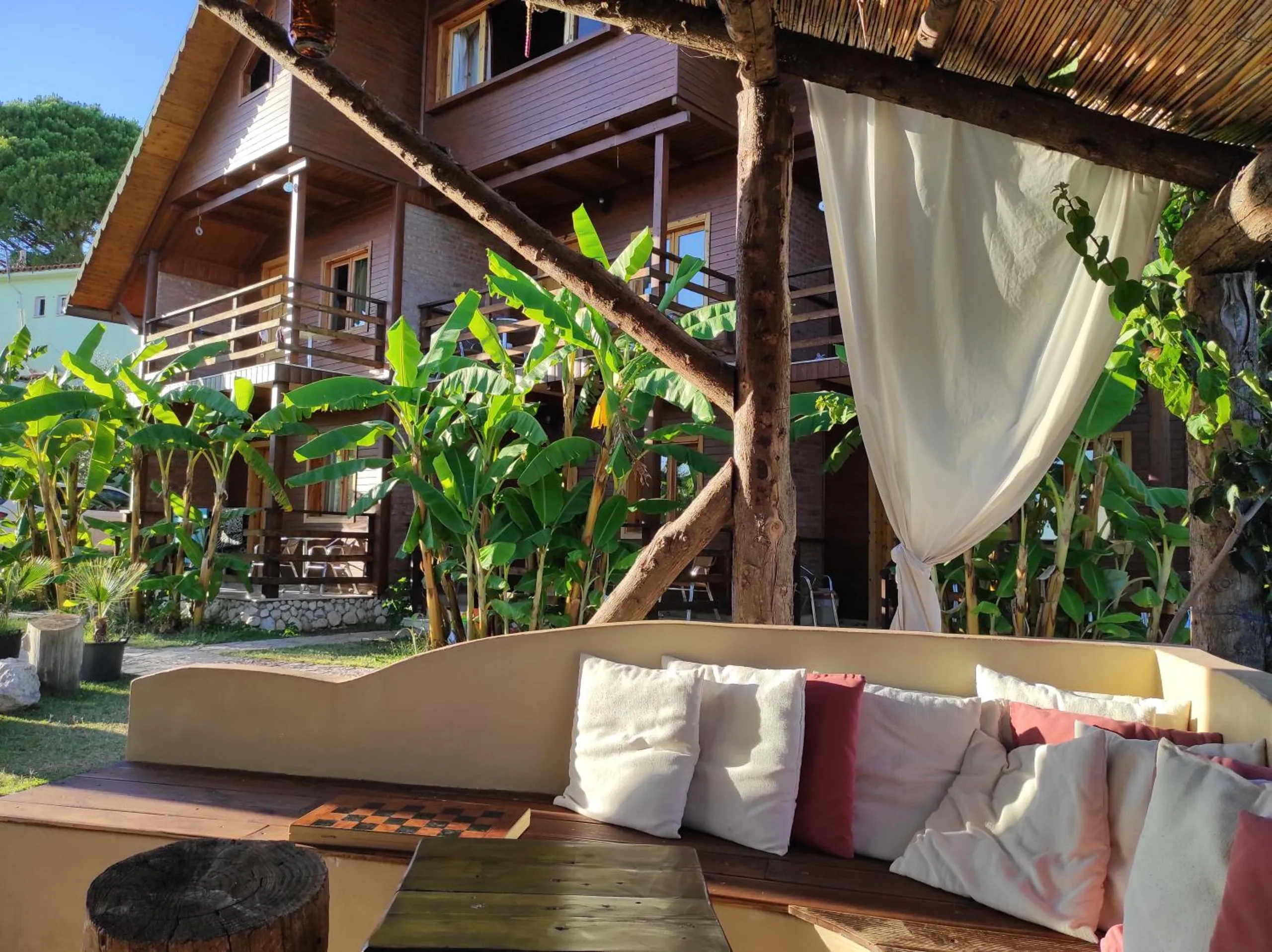 The pavilion lounge by day: a curved rendered banquette with white and red cushions, a chessboard and log stools, the timber chalet and banana plants beyond a tied-back curtain