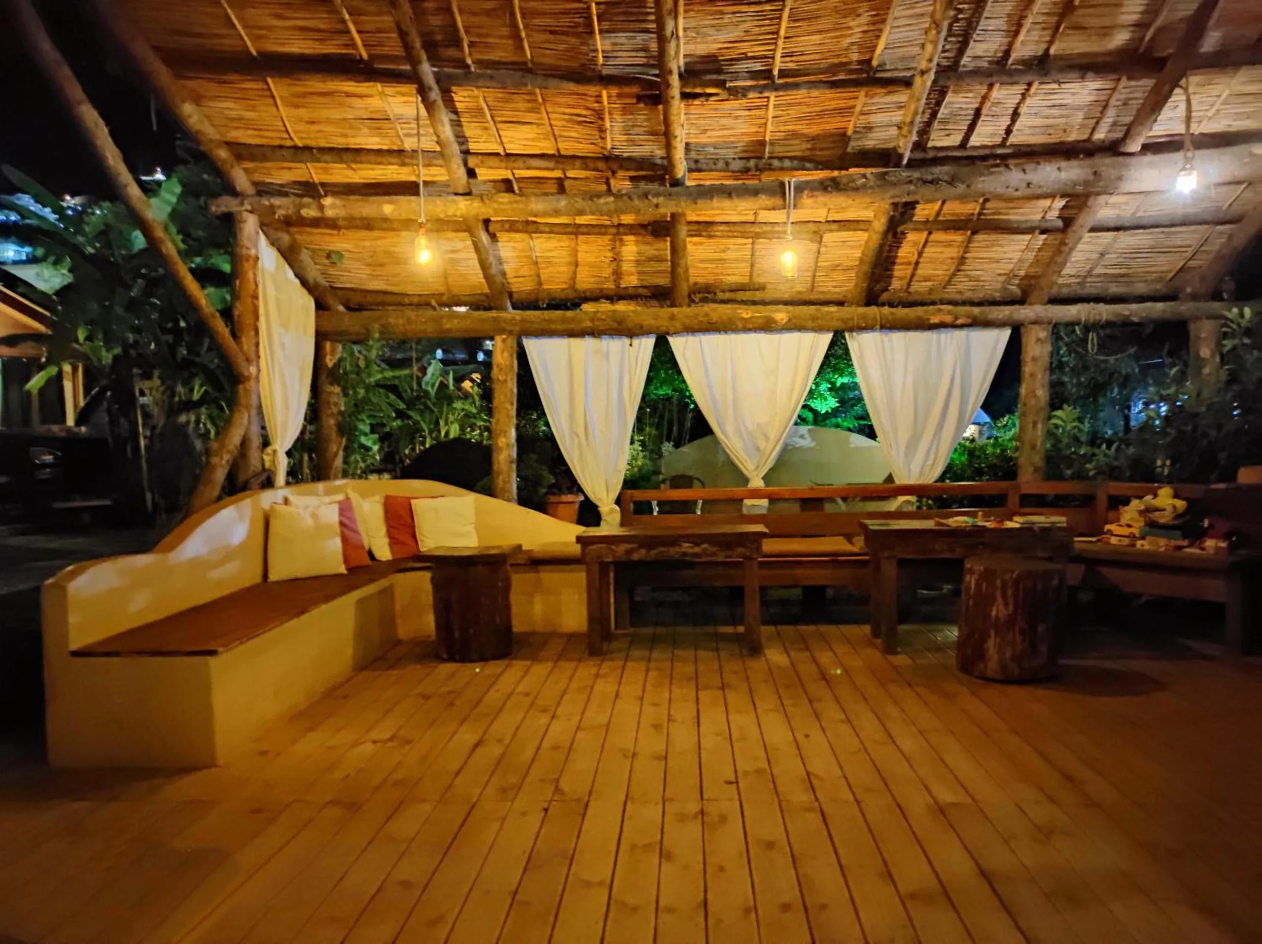 Inside the pavilion at night: a plank floor, a long rendered banquette with cushions, timber tables and white curtains under a reed ceiling hung with bulbs