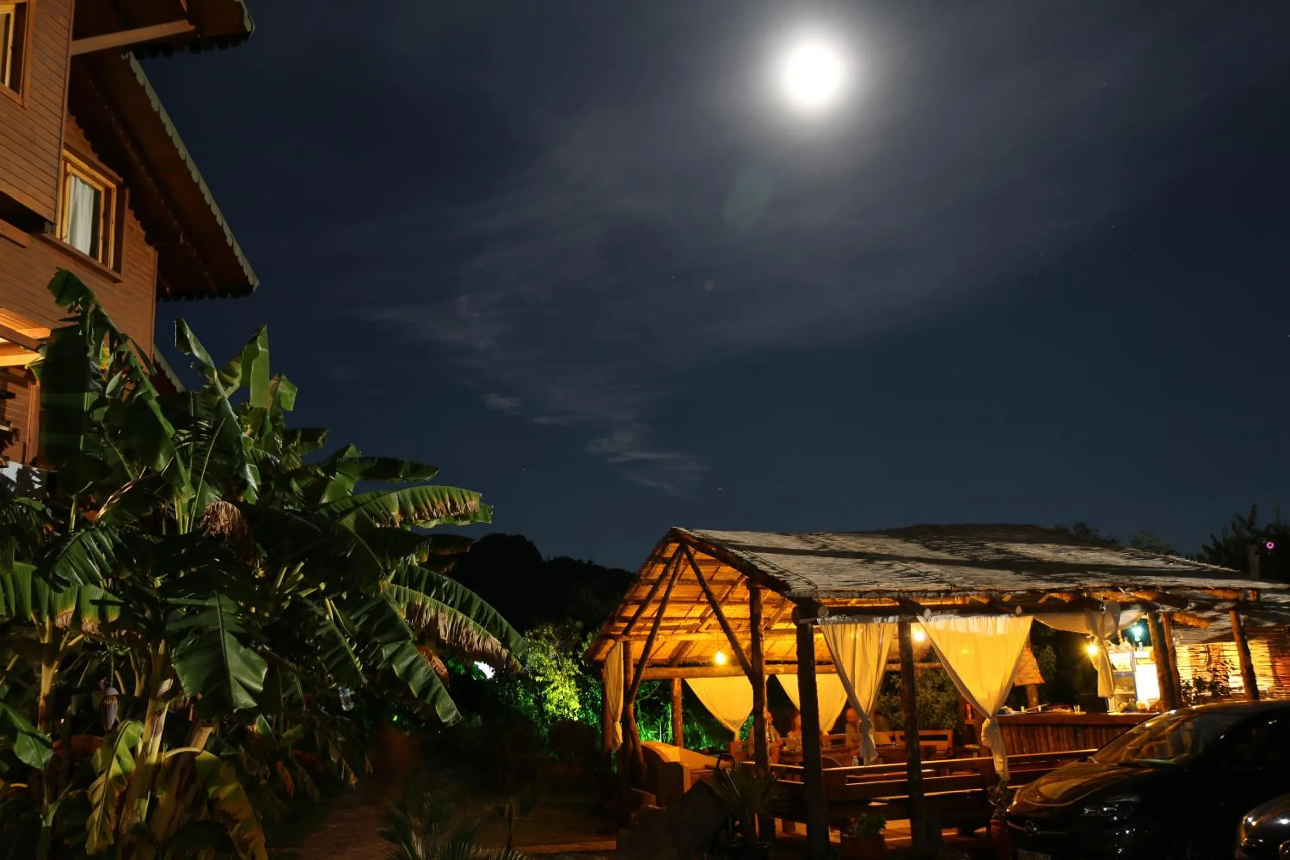 A full moon over the garden at night: banana plants and the corner of a timber chalet on the left, the thatched pavilion lit gold behind white curtains on the right