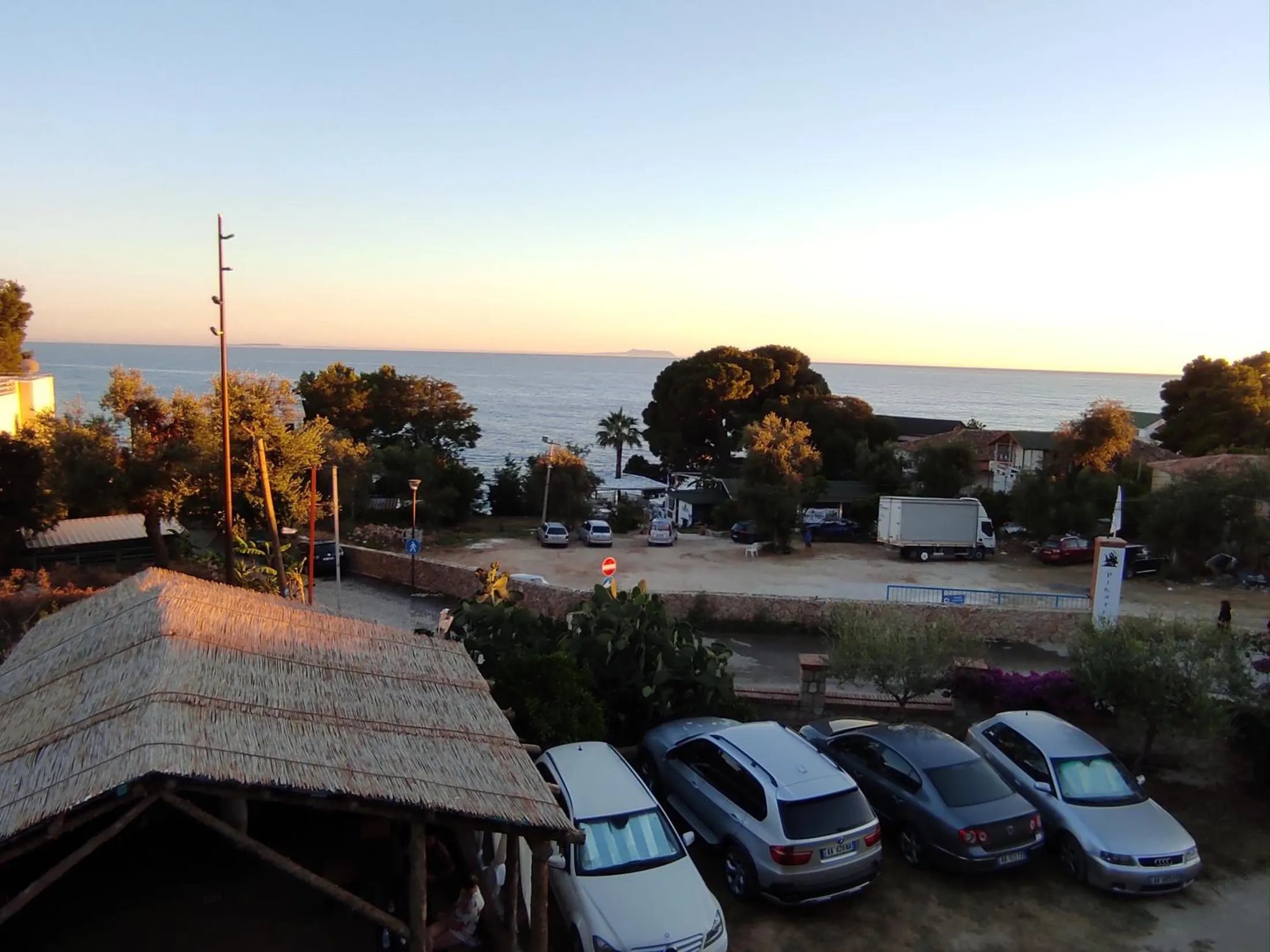 Evening from an upper balcony: the pavilion's thatch and parked cars below, trees, and the sea on the horizon