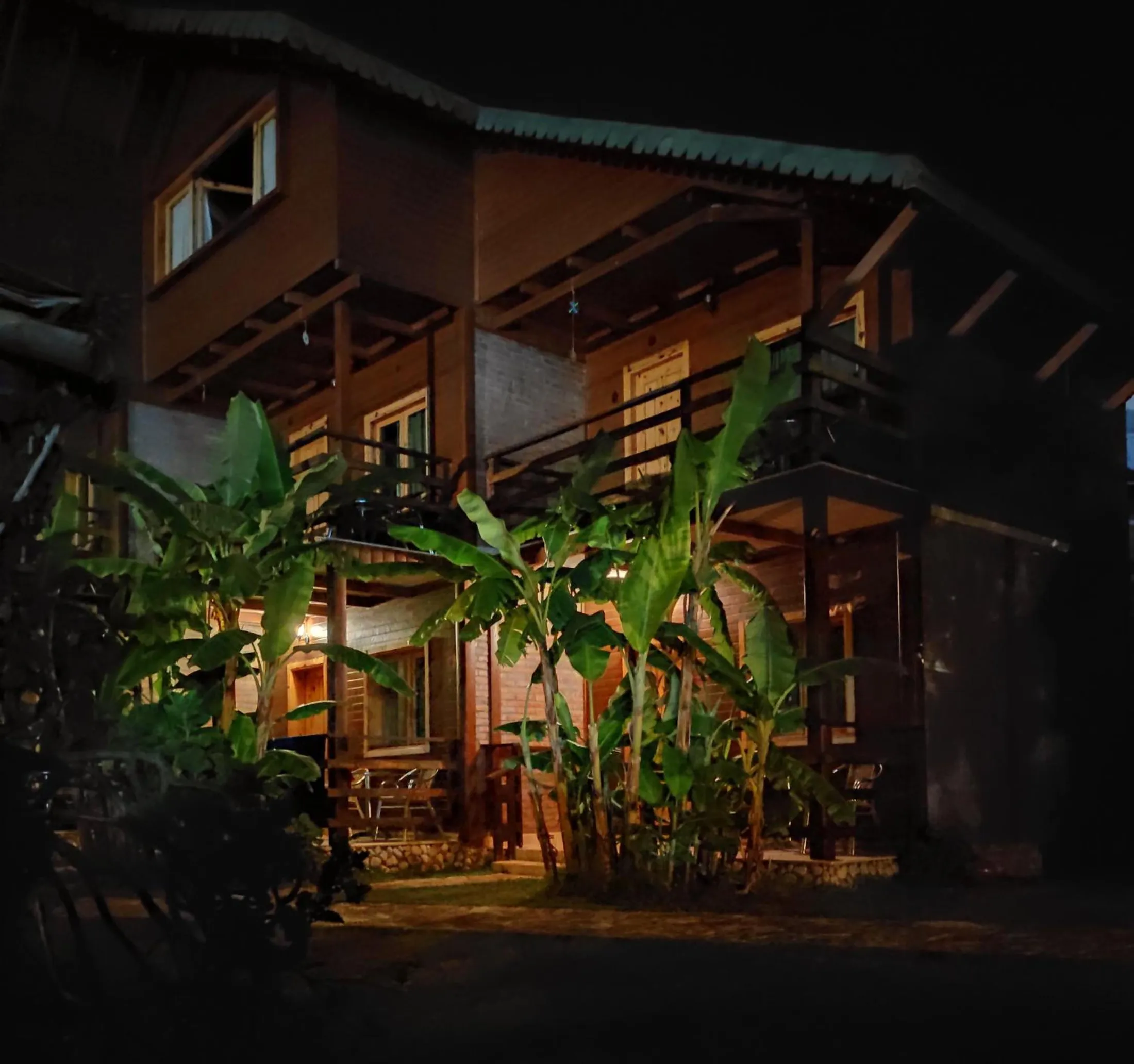 The two-storey timber chalet at night with lit balconies above banana plants