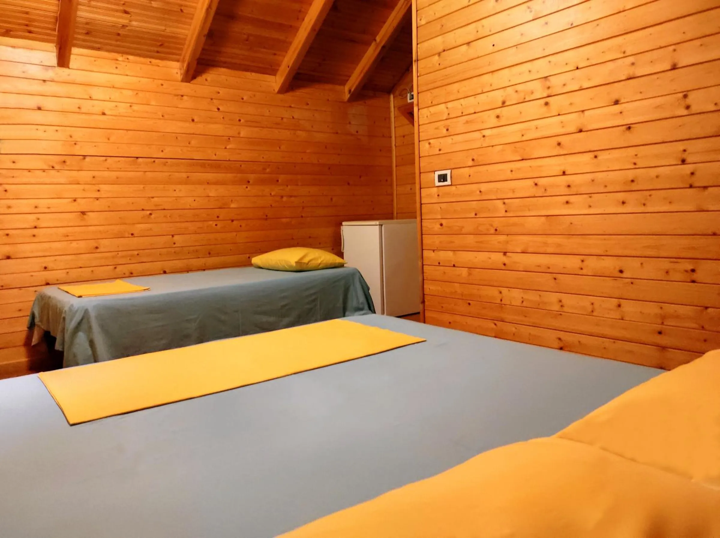 Beds with grey covers and yellow throws in a pine-boarded room under the roof, a small fridge in the corner