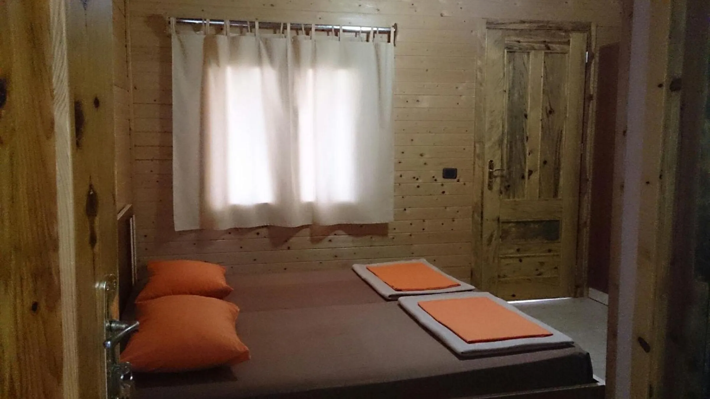 A pine-boarded room with two low beds pushed together under a curtained window, orange cushions and a plank door