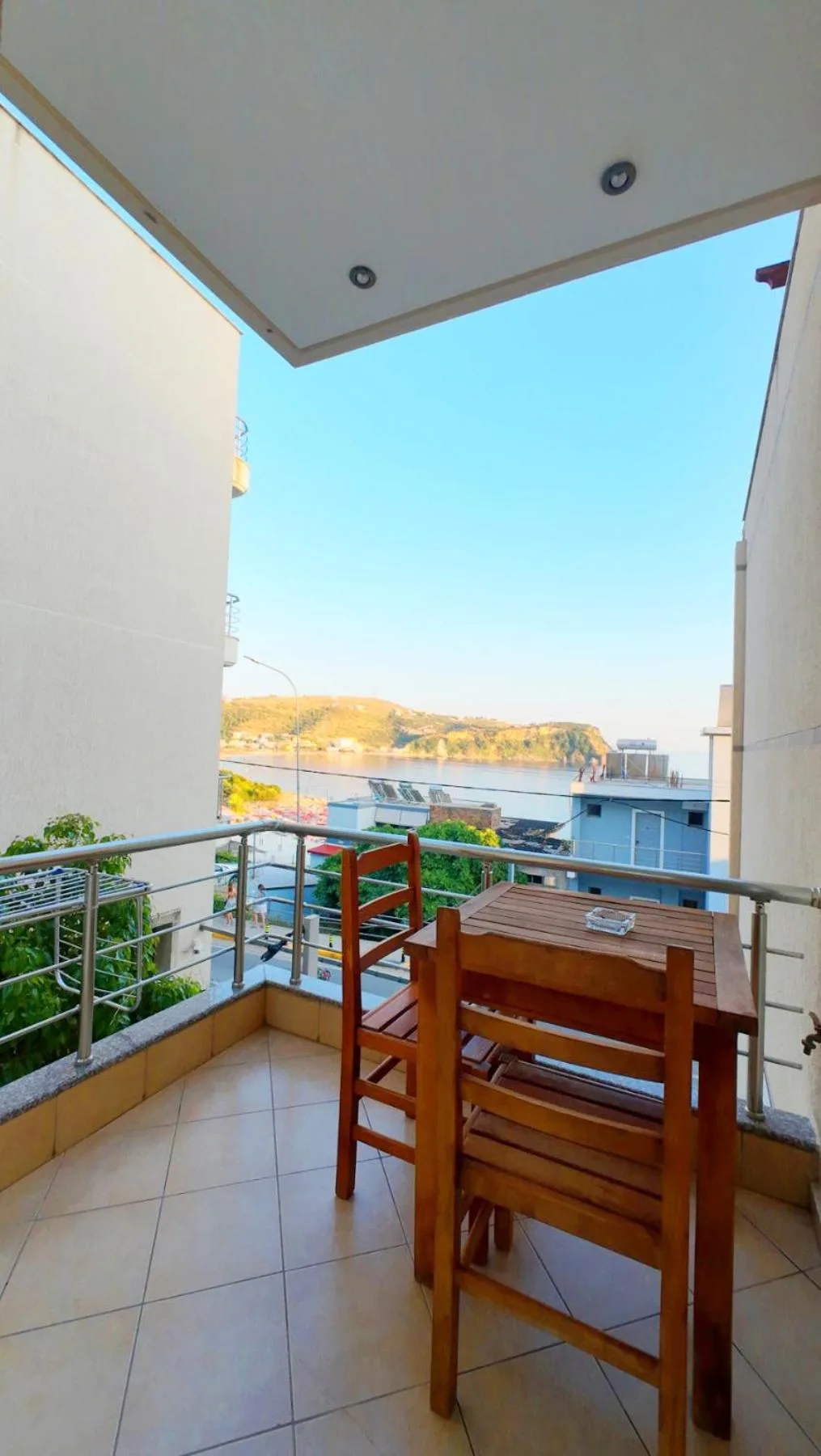 A balcony with a timber table and two chairs, a steel rail, and the bay and the headland beyond