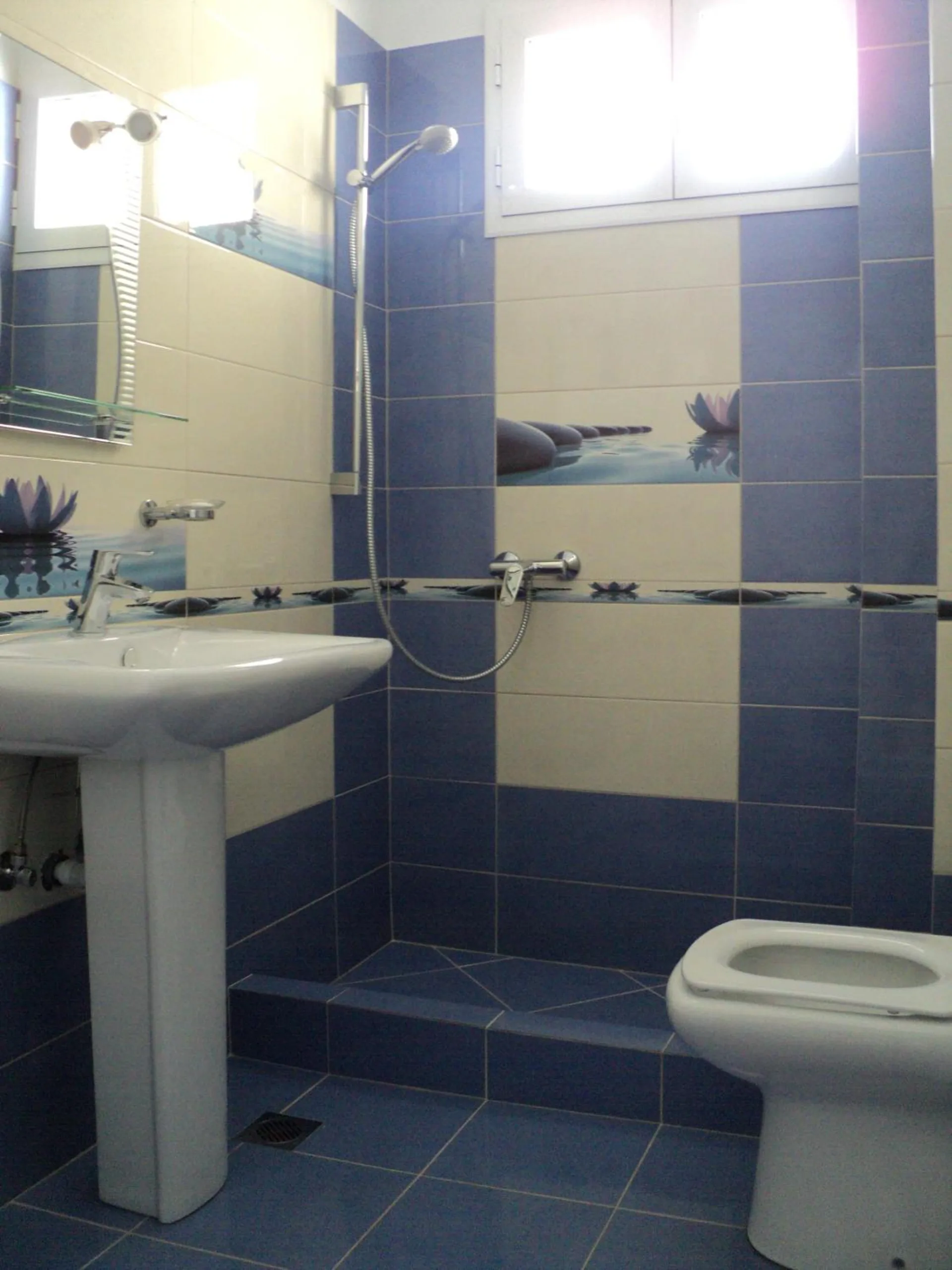 A bathroom tiled cream and blue, with a pedestal basin, a mirror, a step-in shower and a wall-mounted shower head