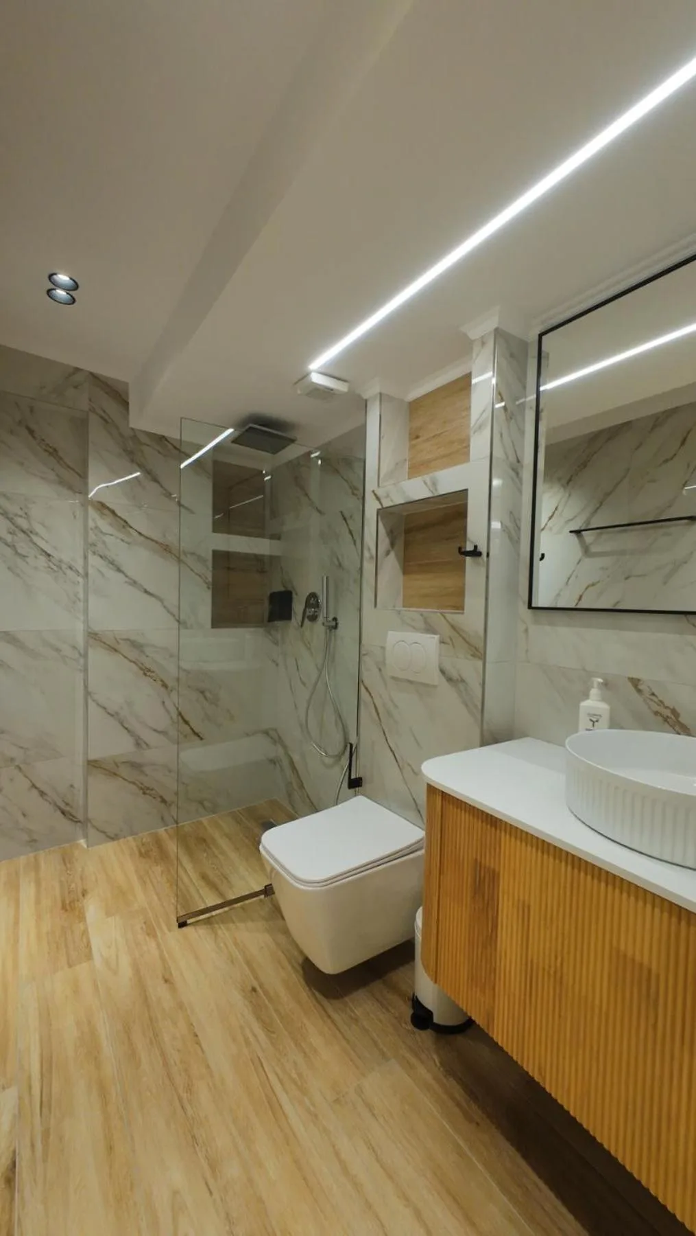 A bathroom with marble-effect walls, a glass shower, a wall-hung toilet, a vessel basin on a ribbed oak vanity and a strip-lit mirror
