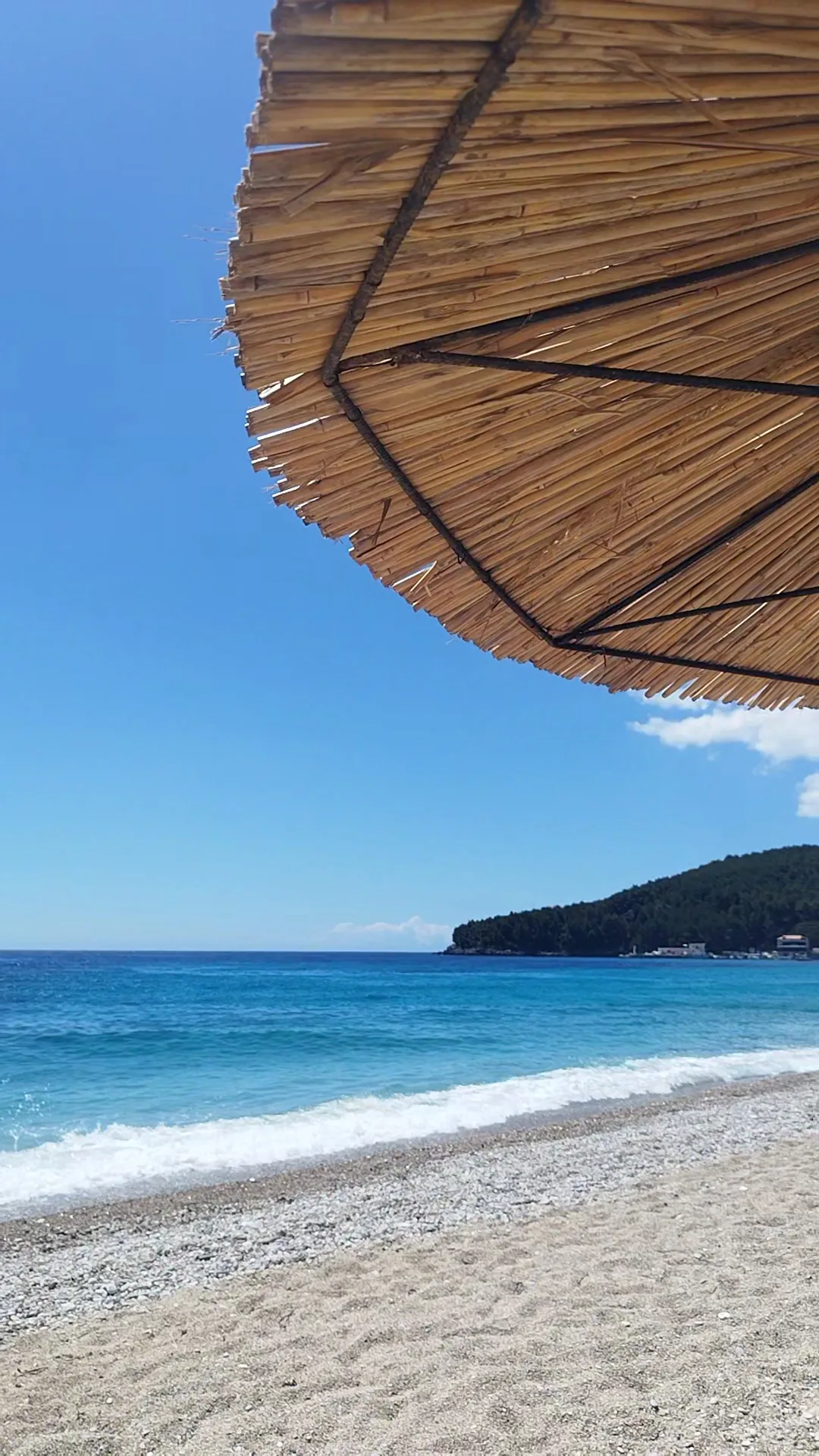 A pebble beach under the edge of a straw umbrella, small waves breaking and a wooded headland across the water