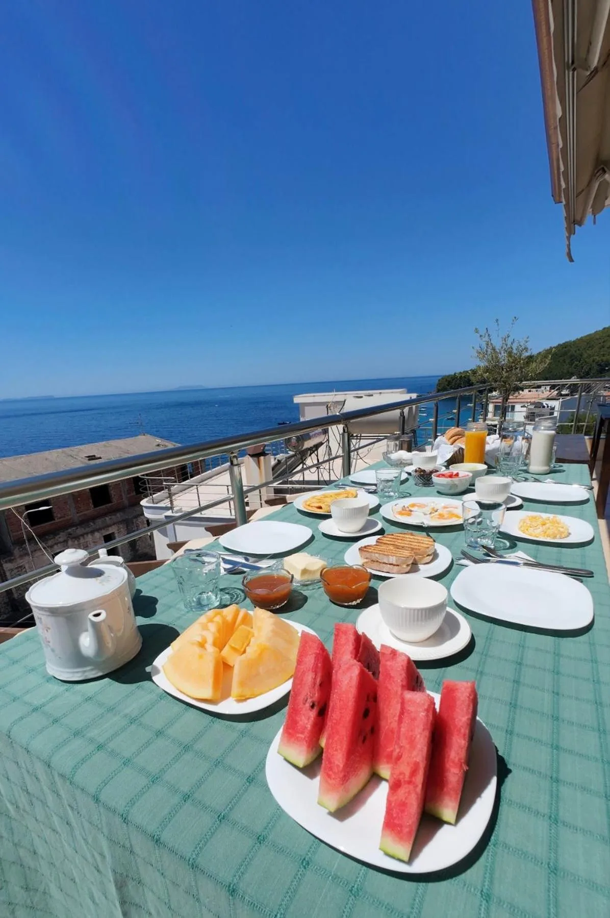 Breakfast laid on a green checked cloth at the terrace rail: watermelon, melon, eggs, bread, jams, juice and a teapot, with the sea and the hills behind
