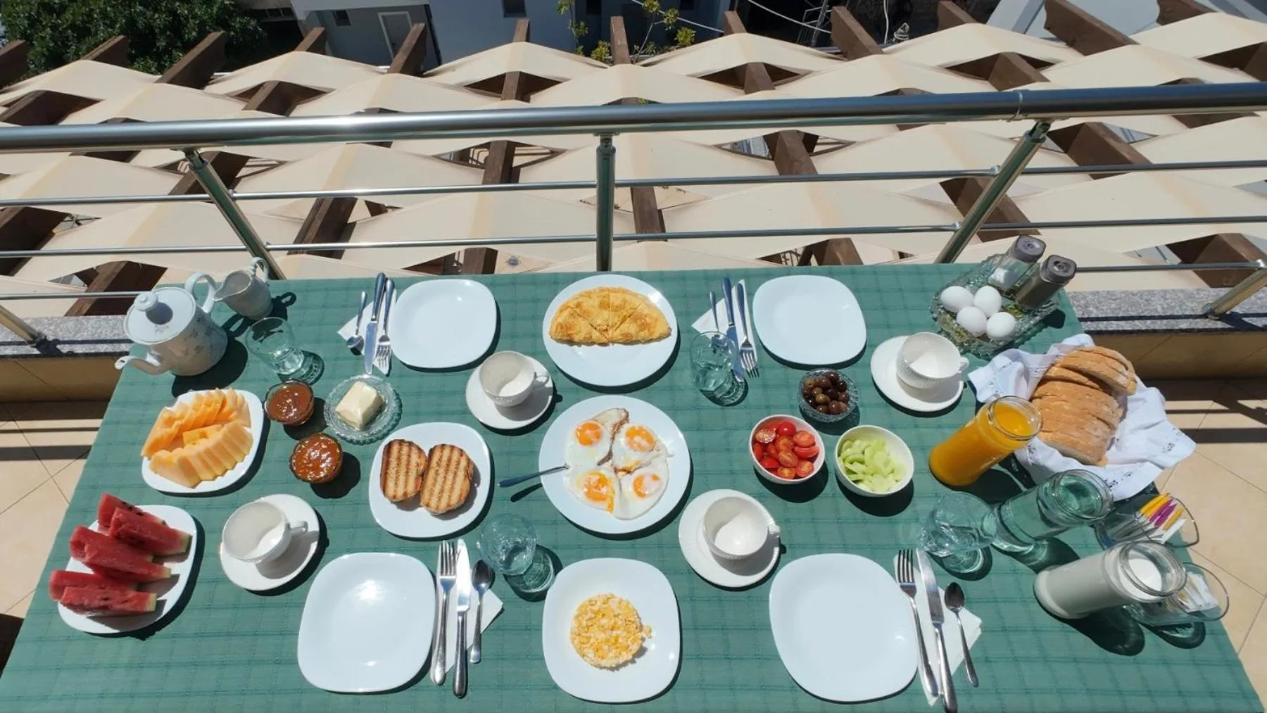A breakfast table seen from above: plates of eggs, bread, tomatoes, watermelon, cheese and fruit on a green checked cloth, with the pergola's woven shade at the top of the frame