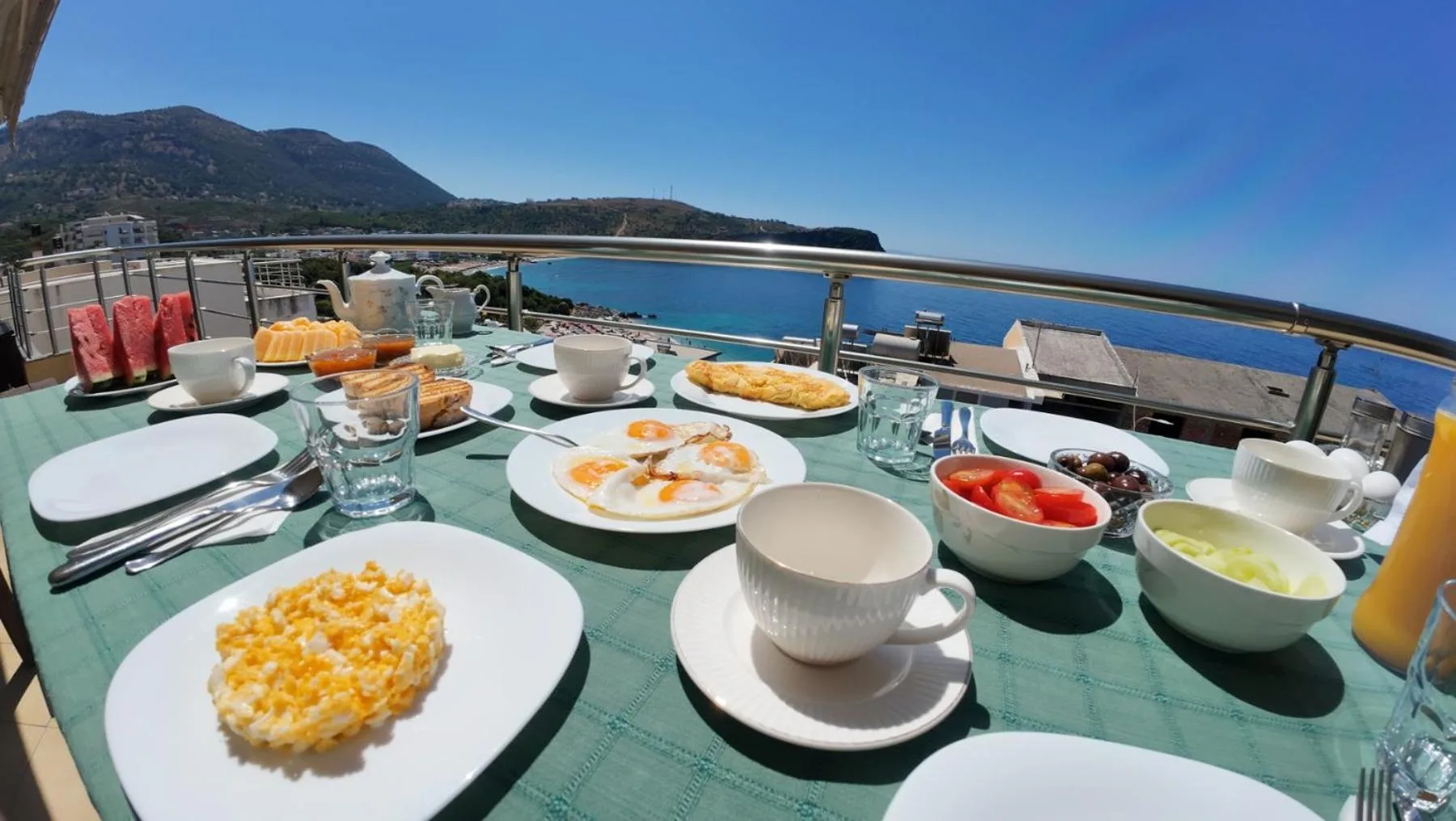 A breakfast table at the terrace rail with scrambled eggs, fried eggs, tomatoes, coffee and juice, the bay and its hills beyond