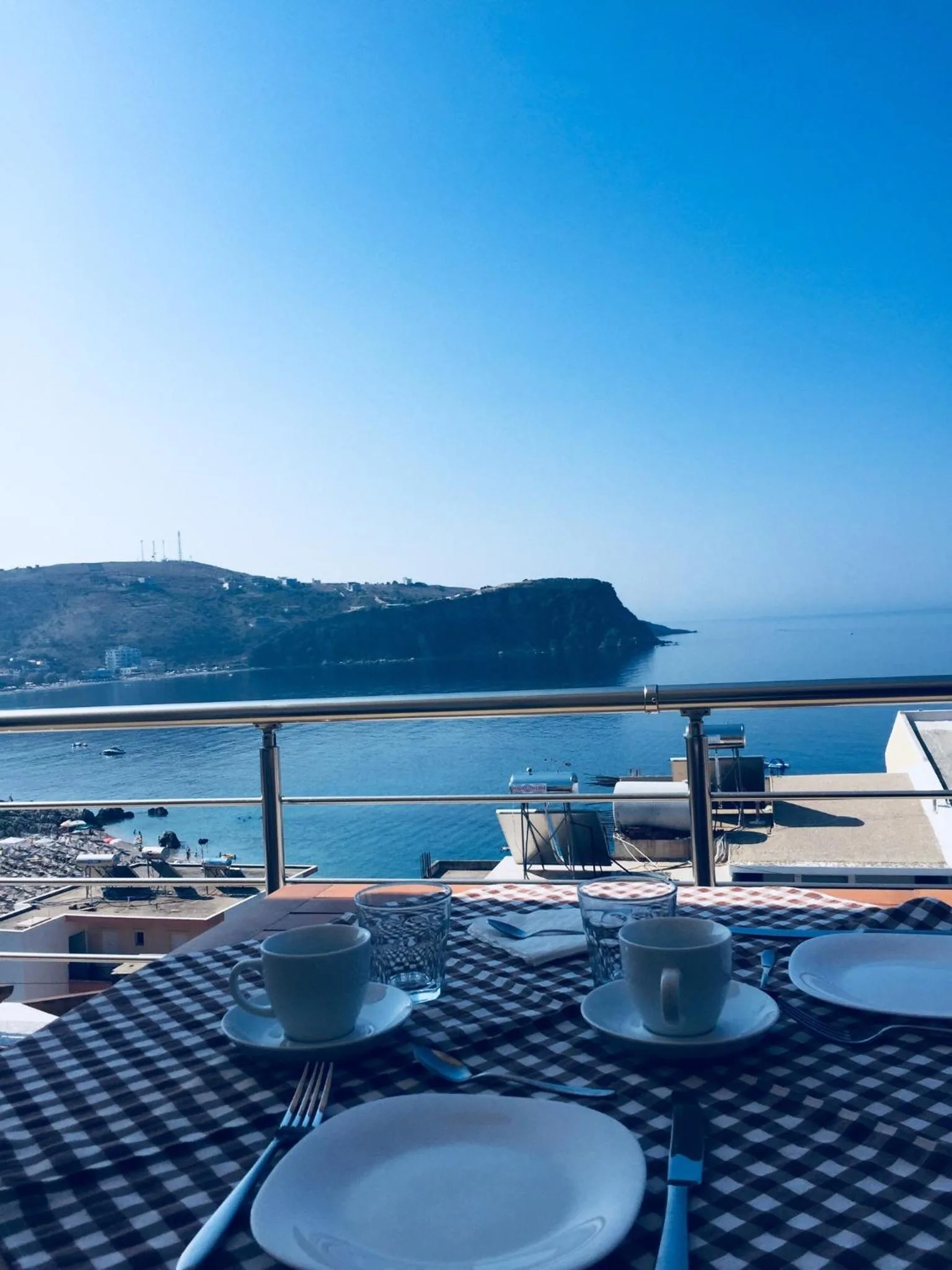 Two coffee cups and plates on a blue checked tablecloth at the terrace rail, with the rocky headland and the sea beyond under a clear sky