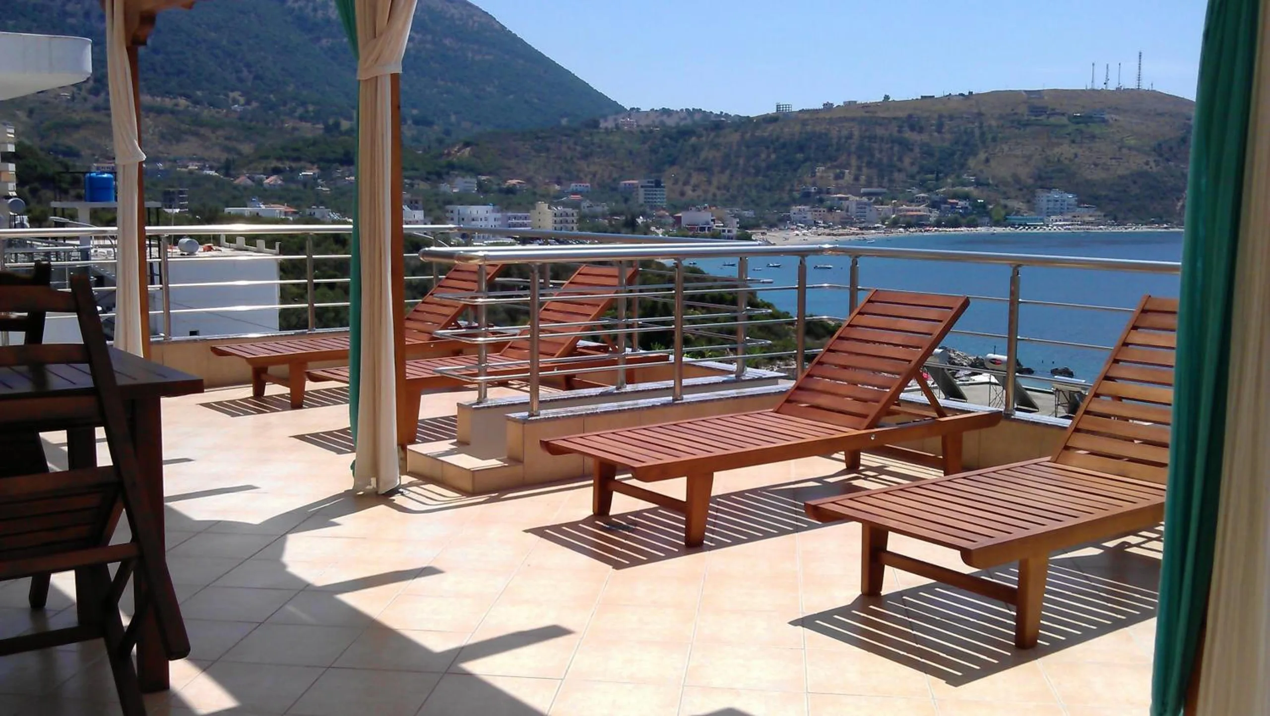 Wooden loungers on the sunlit roof terrace, a table and chairs to one side, and the bay with the hills behind Himarë beyond the rail