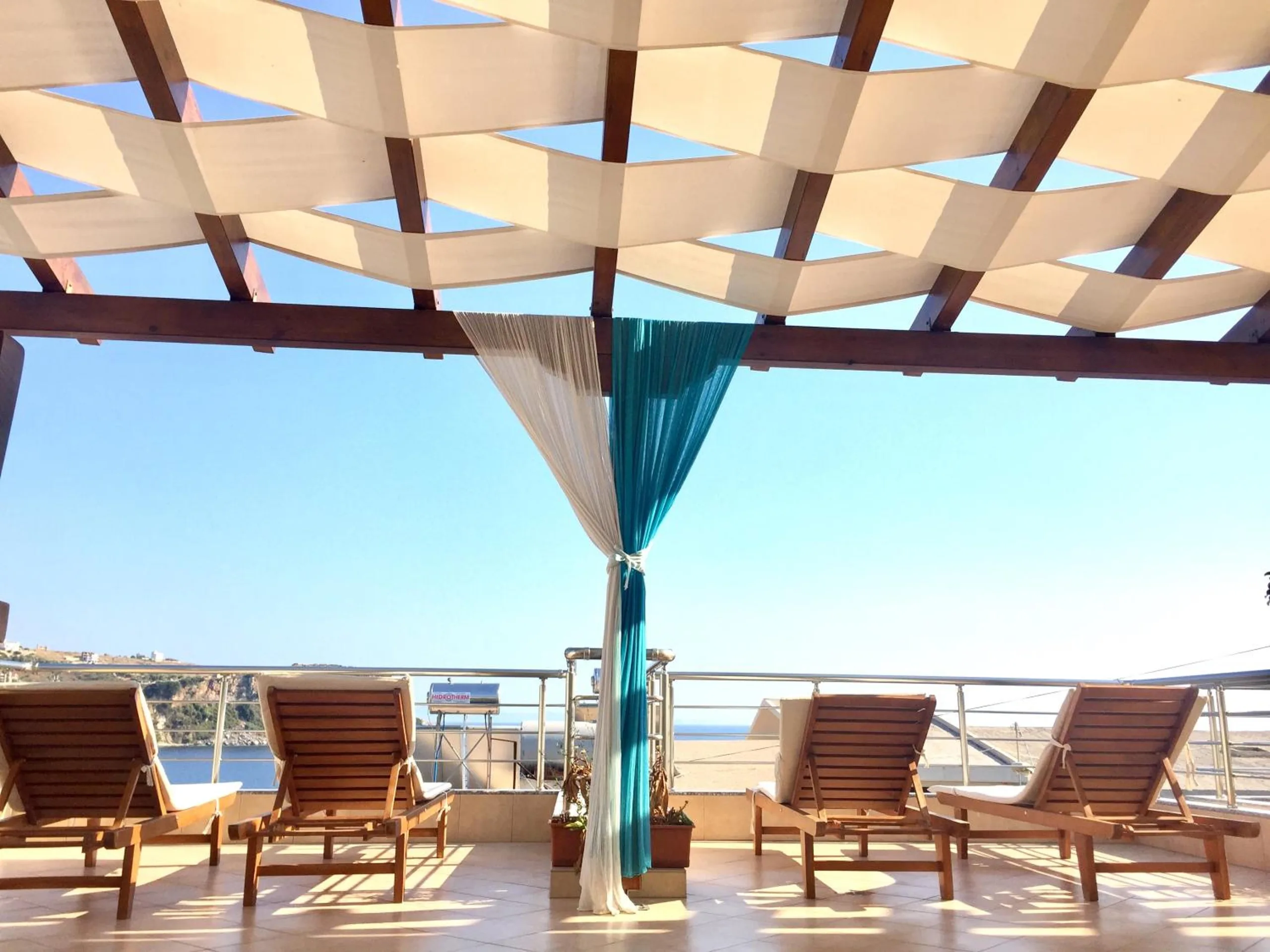 The roof terrace under the woven canvas pergola: four wooden loungers in a row, a teal and ivory drape tied to a post, and the sea beyond the rail