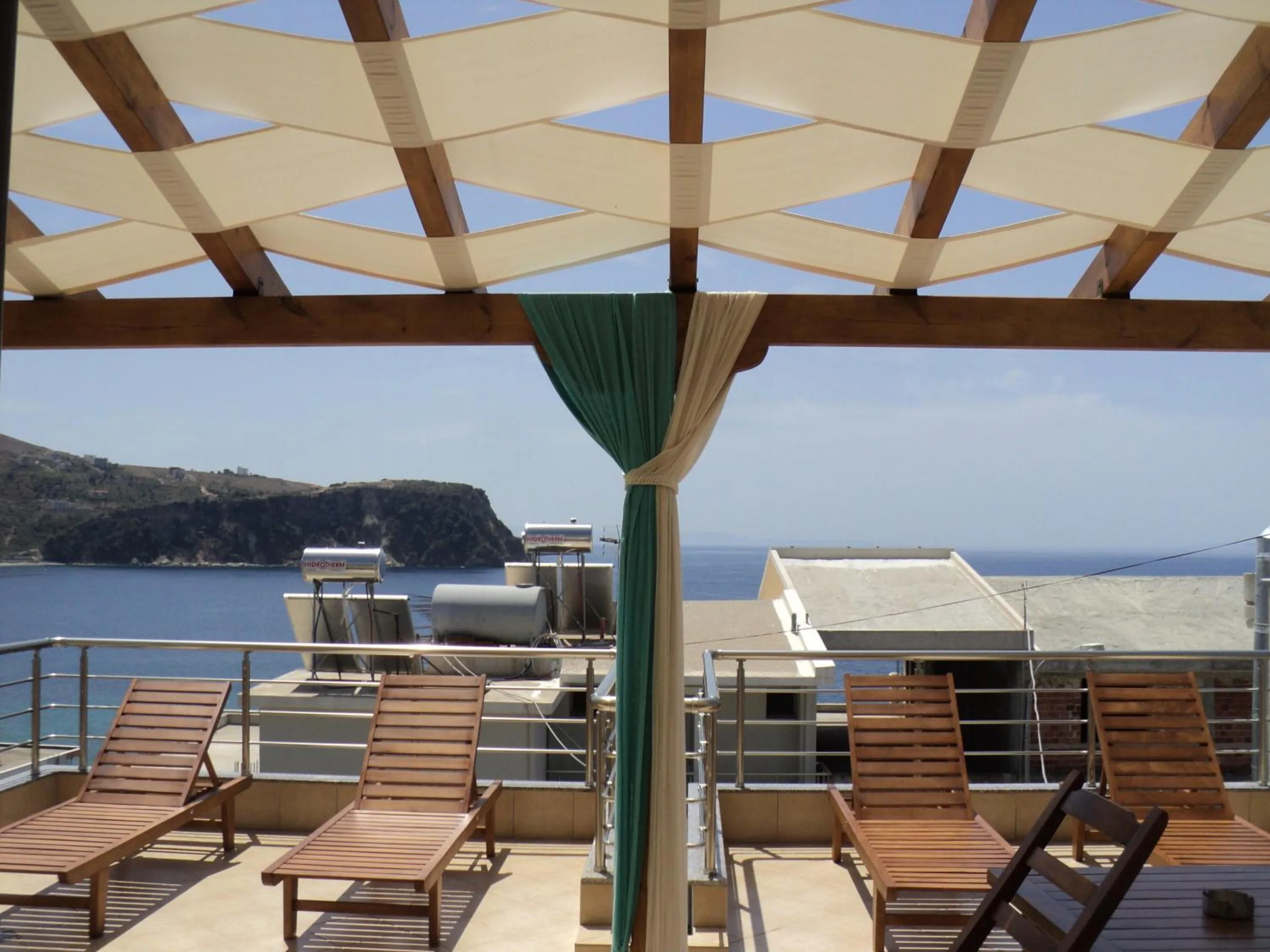 The pergola's woven canvas and timber beams over wooden loungers, with neighbouring rooftops, the sea and the headland beyond
