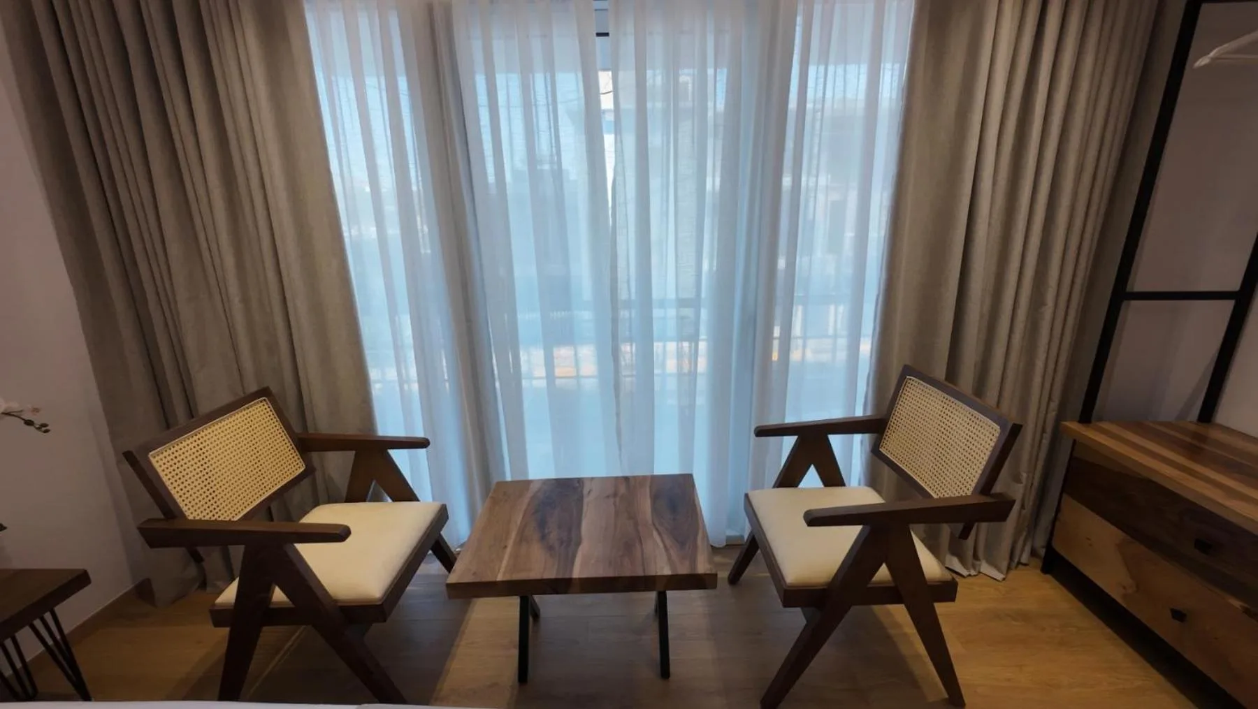 Two cane-backed timber armchairs and a walnut table in front of sheer white curtains and a balcony door, in one of the newer rooms