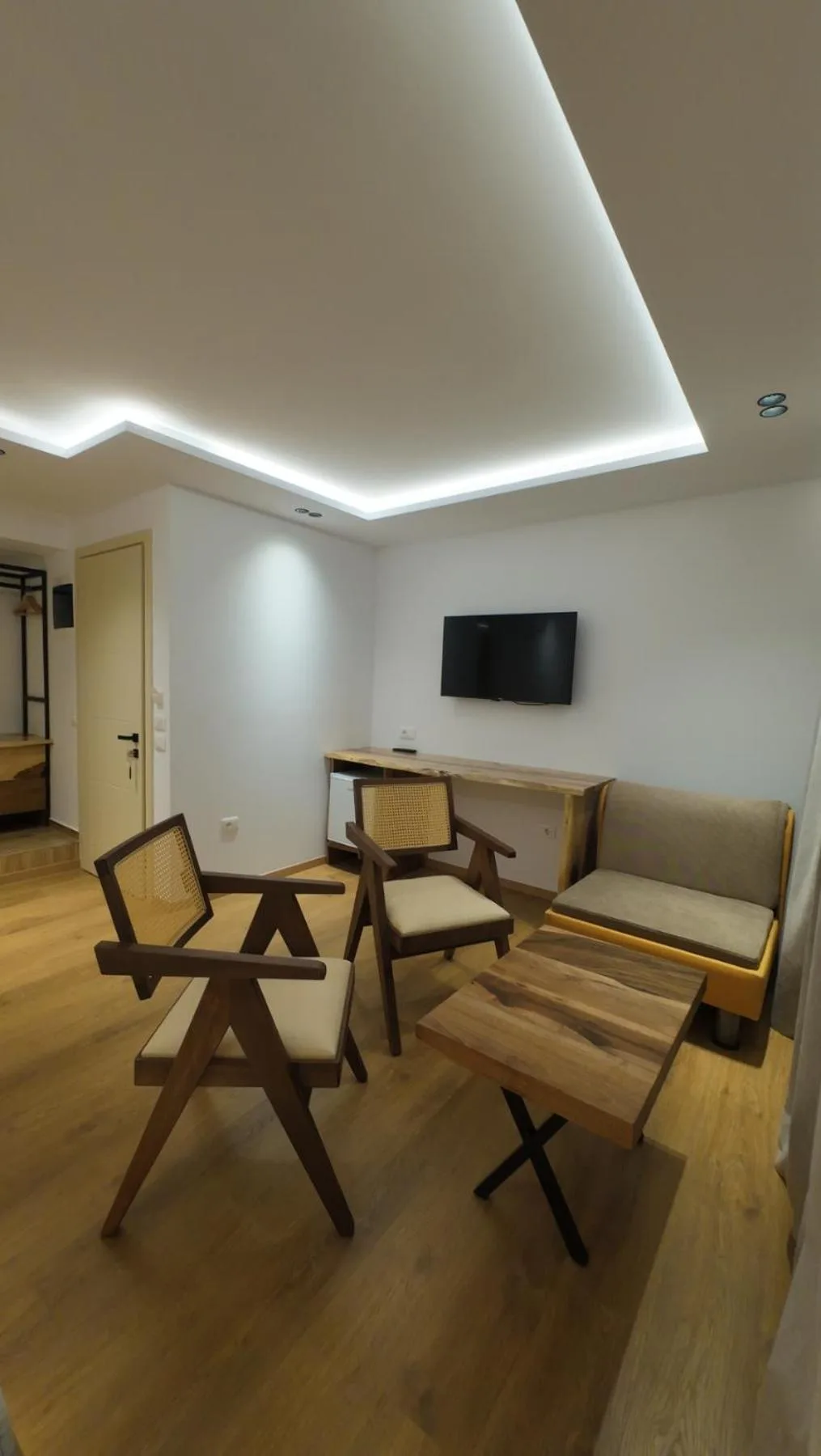 A seating corner with cane armchairs, a timber coffee table and a sofa, a wall-mounted TV and a lit cove ceiling