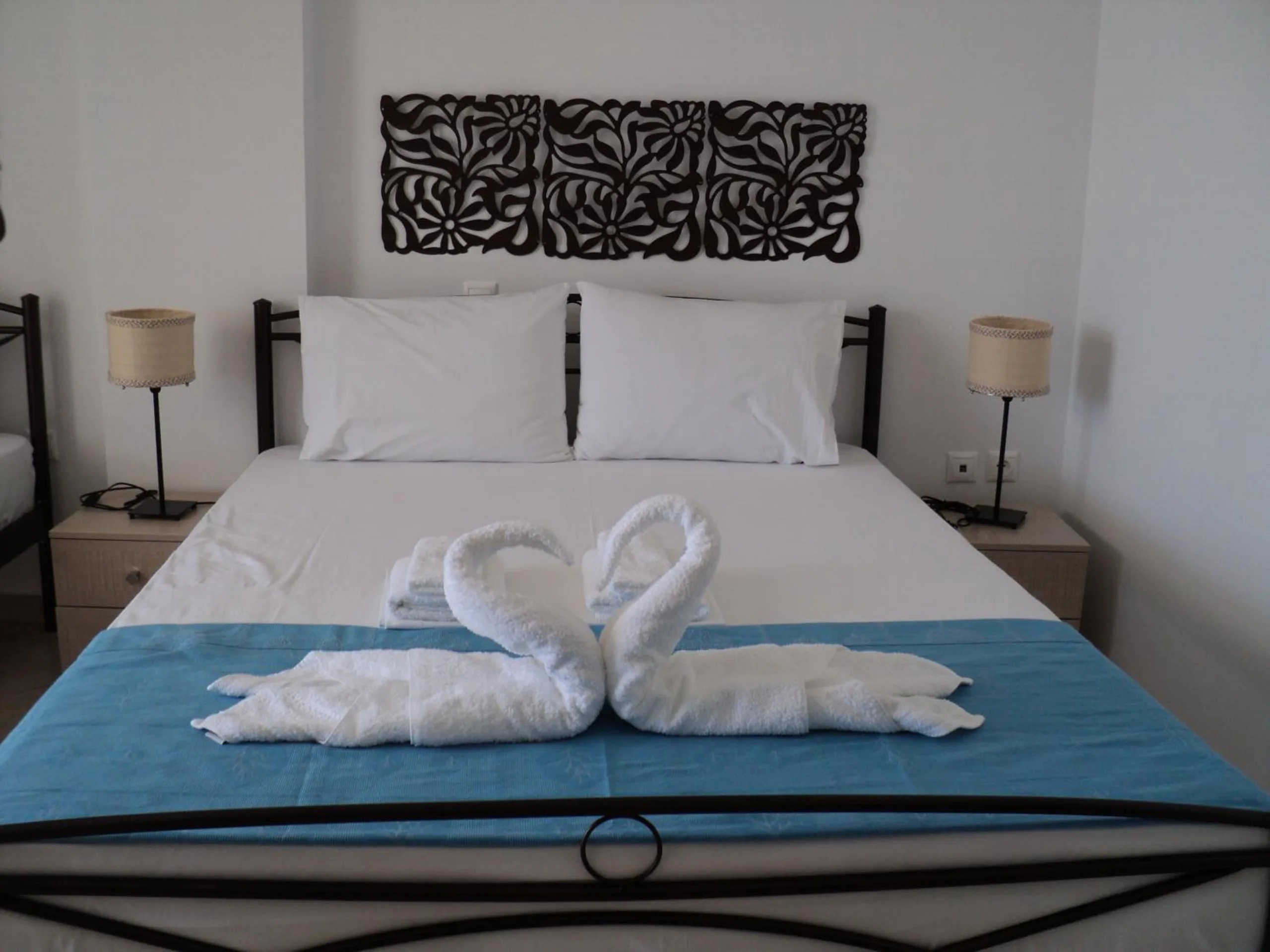 A double bed with a wrought-iron frame and a teal throw, two towels folded into swans, a carved dark timber panel above the bed and two bedside lamps
