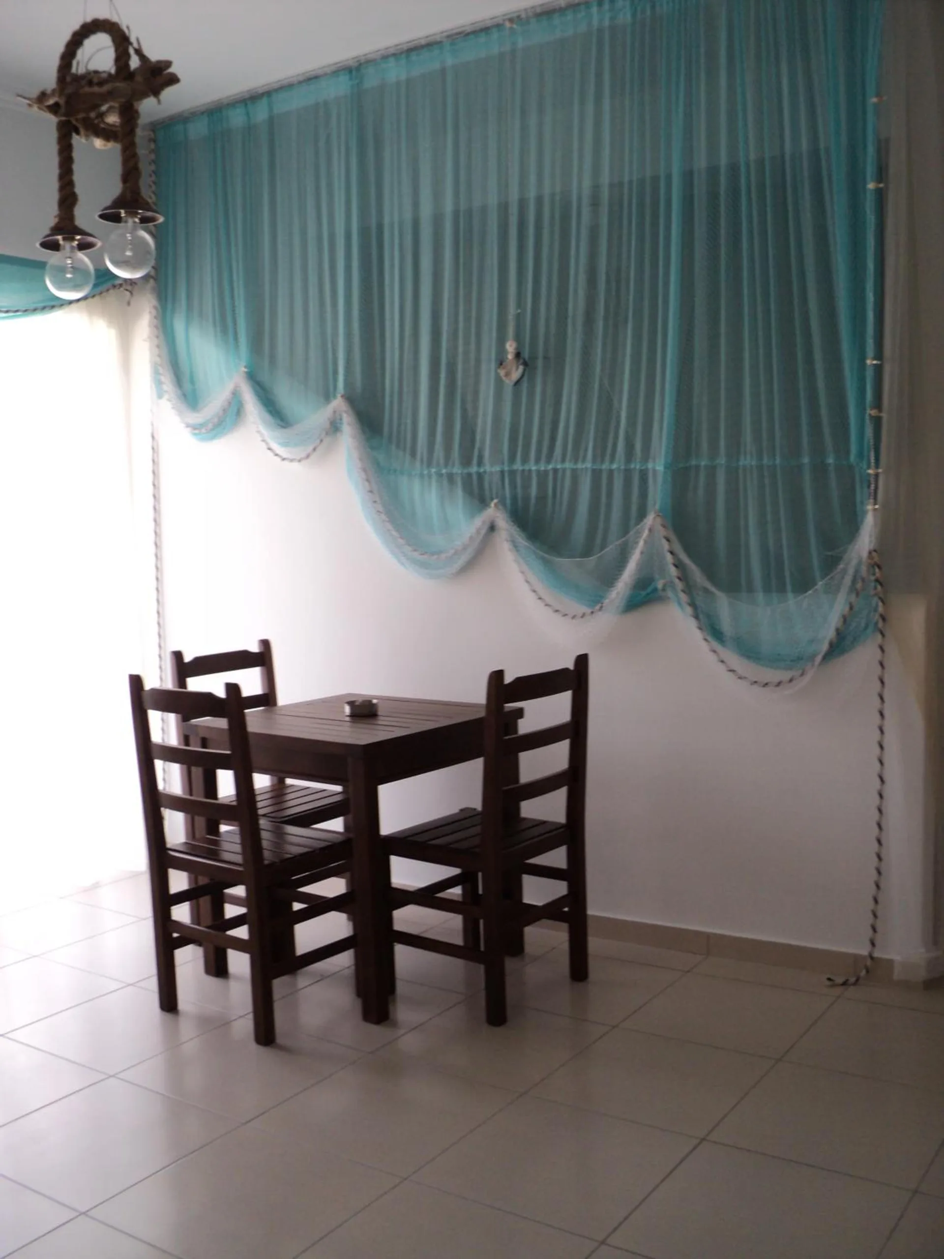 A teal sheer drape gathered in swags over a window, a dark timber table with three chairs below it and a rope-hung pendant light