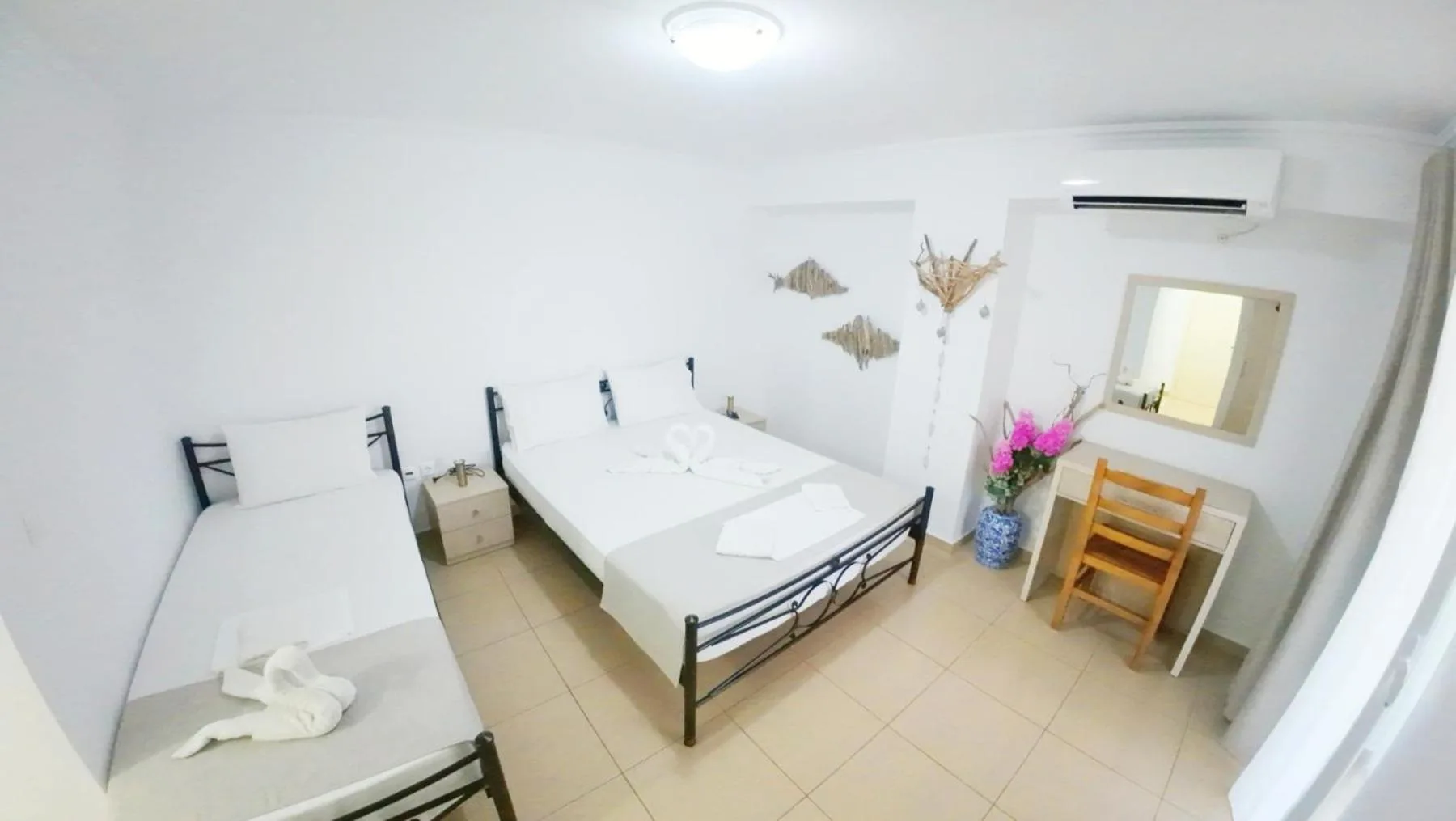 A bright white room with a double and a single wrought-iron bed, a desk with a mirror and a vase of pink flowers, and timber fish on the wall