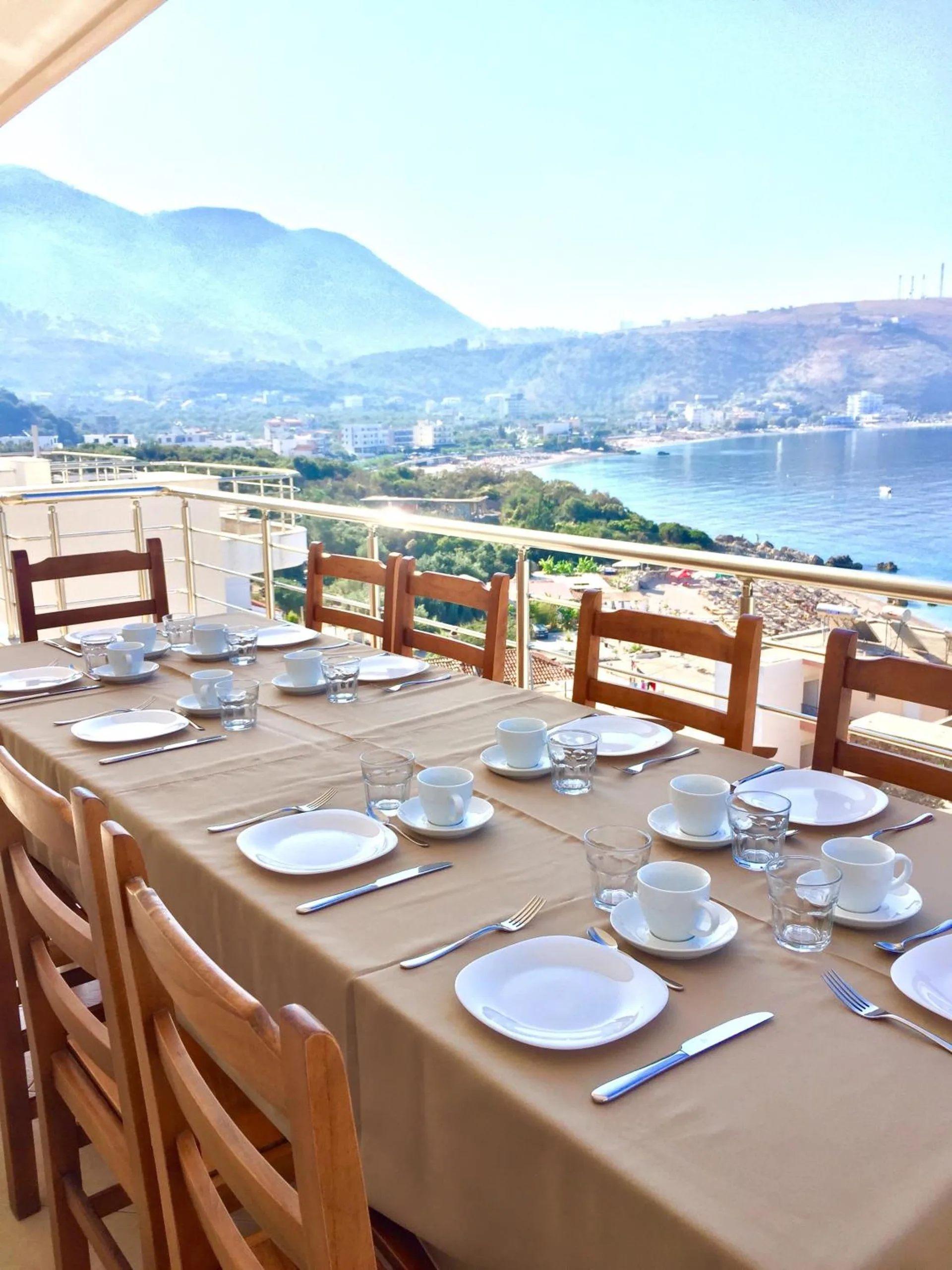 A long table laid for breakfast along the terrace rail with cups, plates and wooden chairs, looking along the coast to the town and the mountains beyond