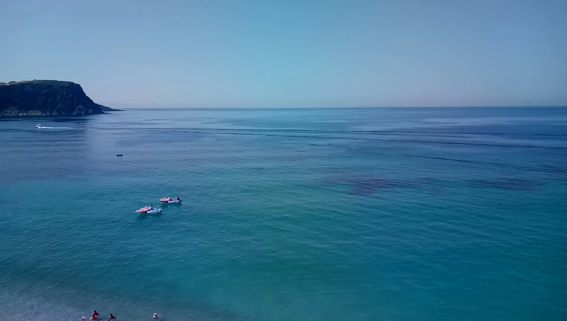 Clear turquoise water with two paddleboarders, a wooded headland on the left and the open sea to the horizon