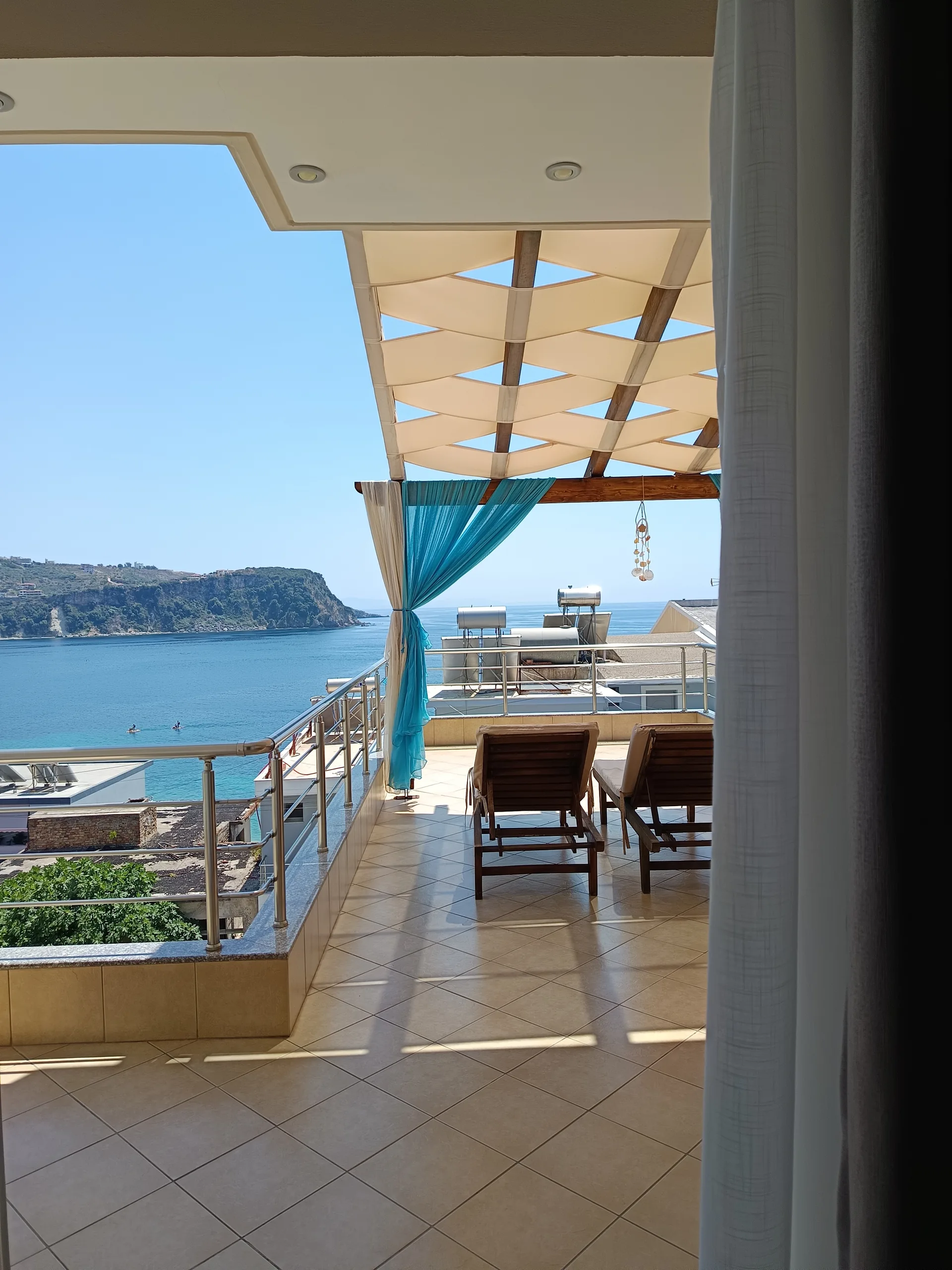 Looking out through a curtained doorway onto the tiled roof terrace of Vila Kosteli: two wooden loungers under a woven cream canvas pergola, a teal drape tied to the post, and the bay with a wooded headland beyond the rail