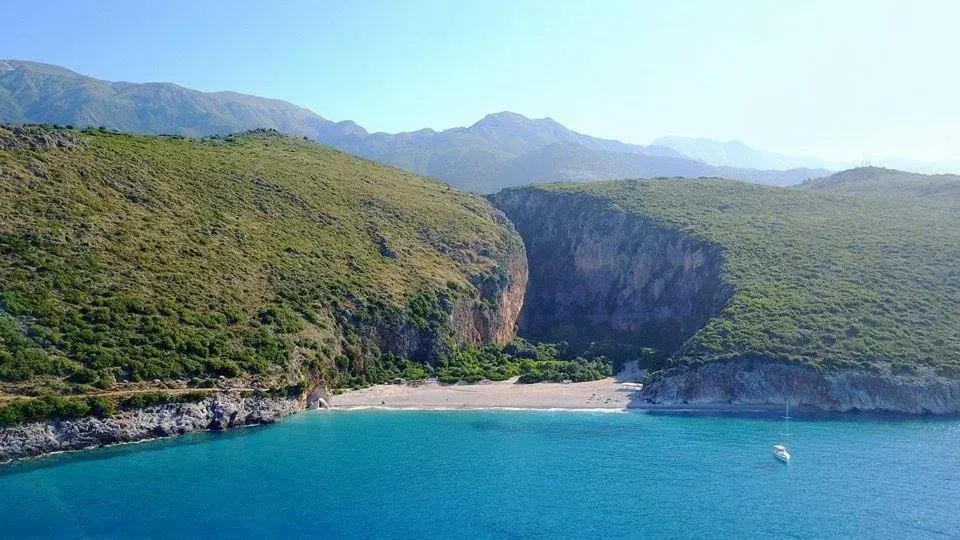 A cove of pale shingle between high cliffs, a boat anchored on turquoise water and a wooded gorge behind