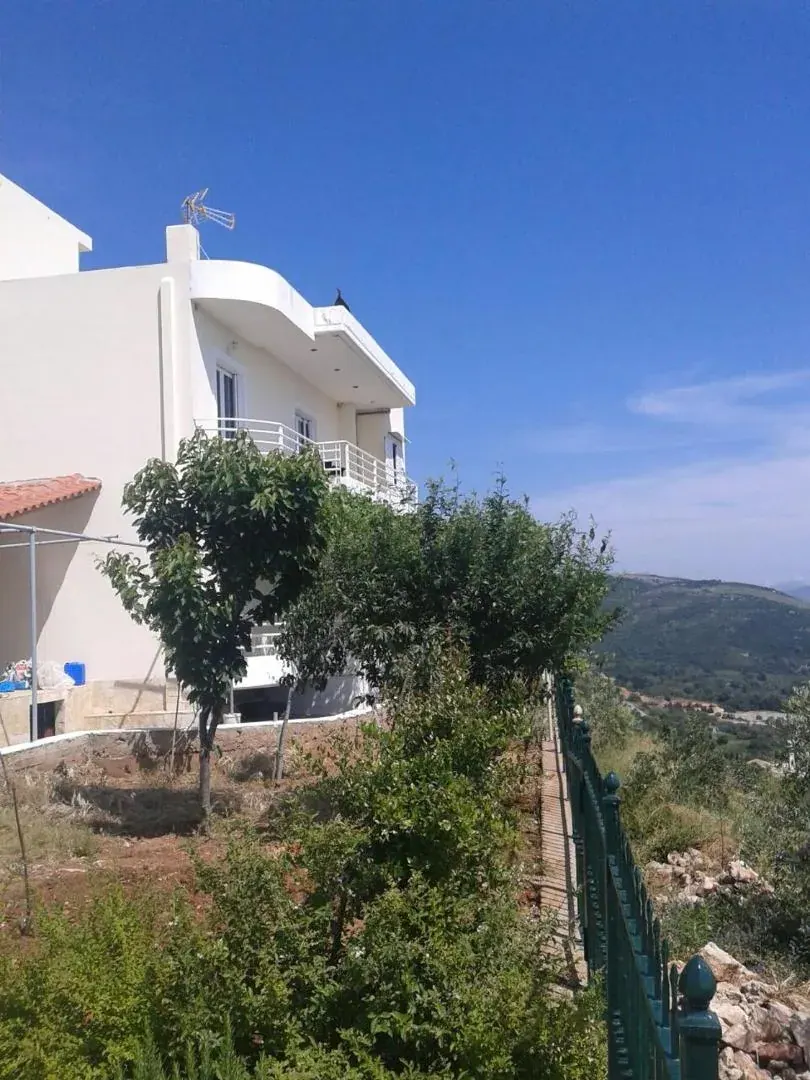 A white three-storey house with railed balconies on a slope, a tree in front of it and hills behind