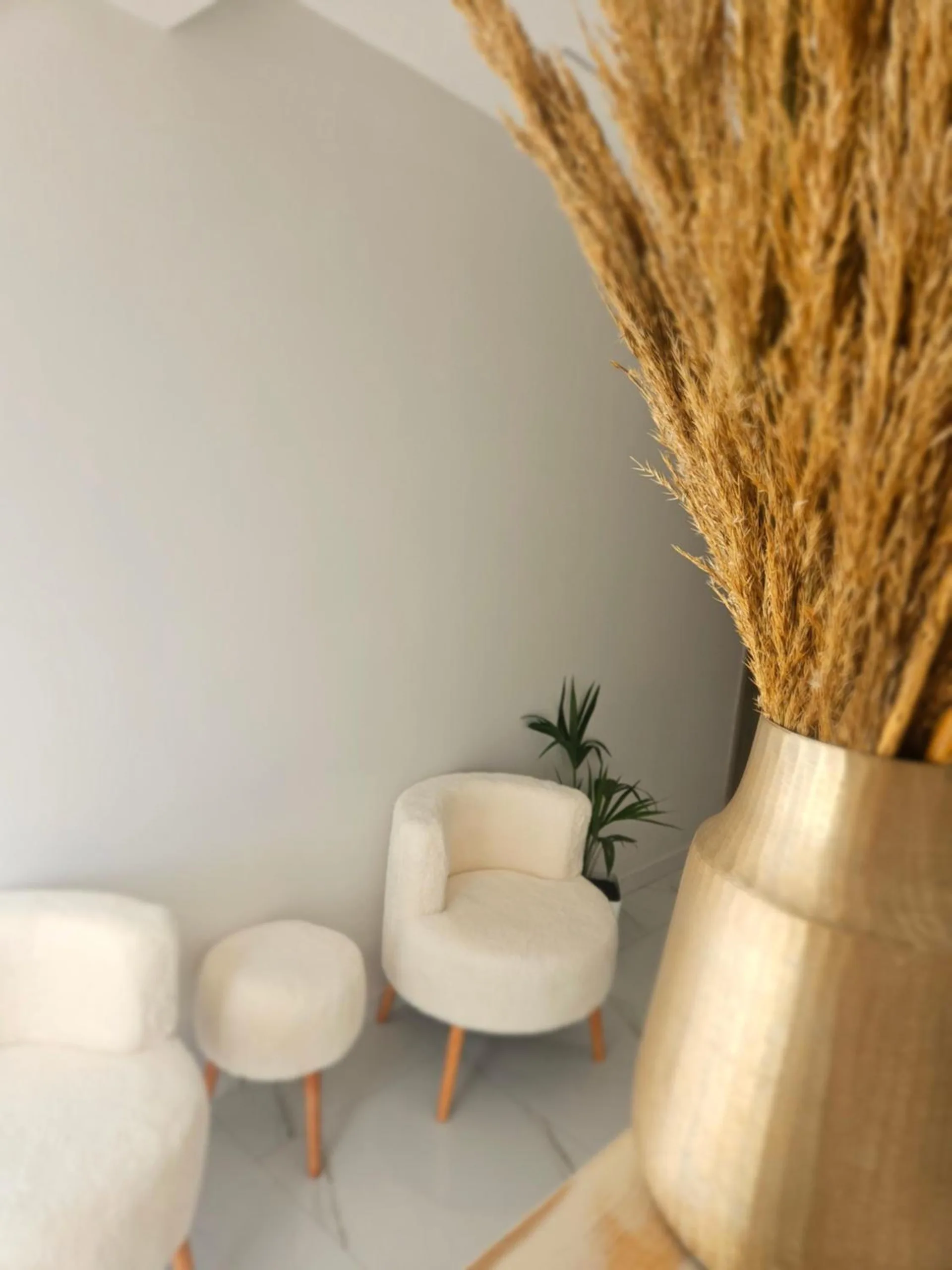Two cream bouclé armchairs and a stool in a white corner, a vase of pampas grass beside them