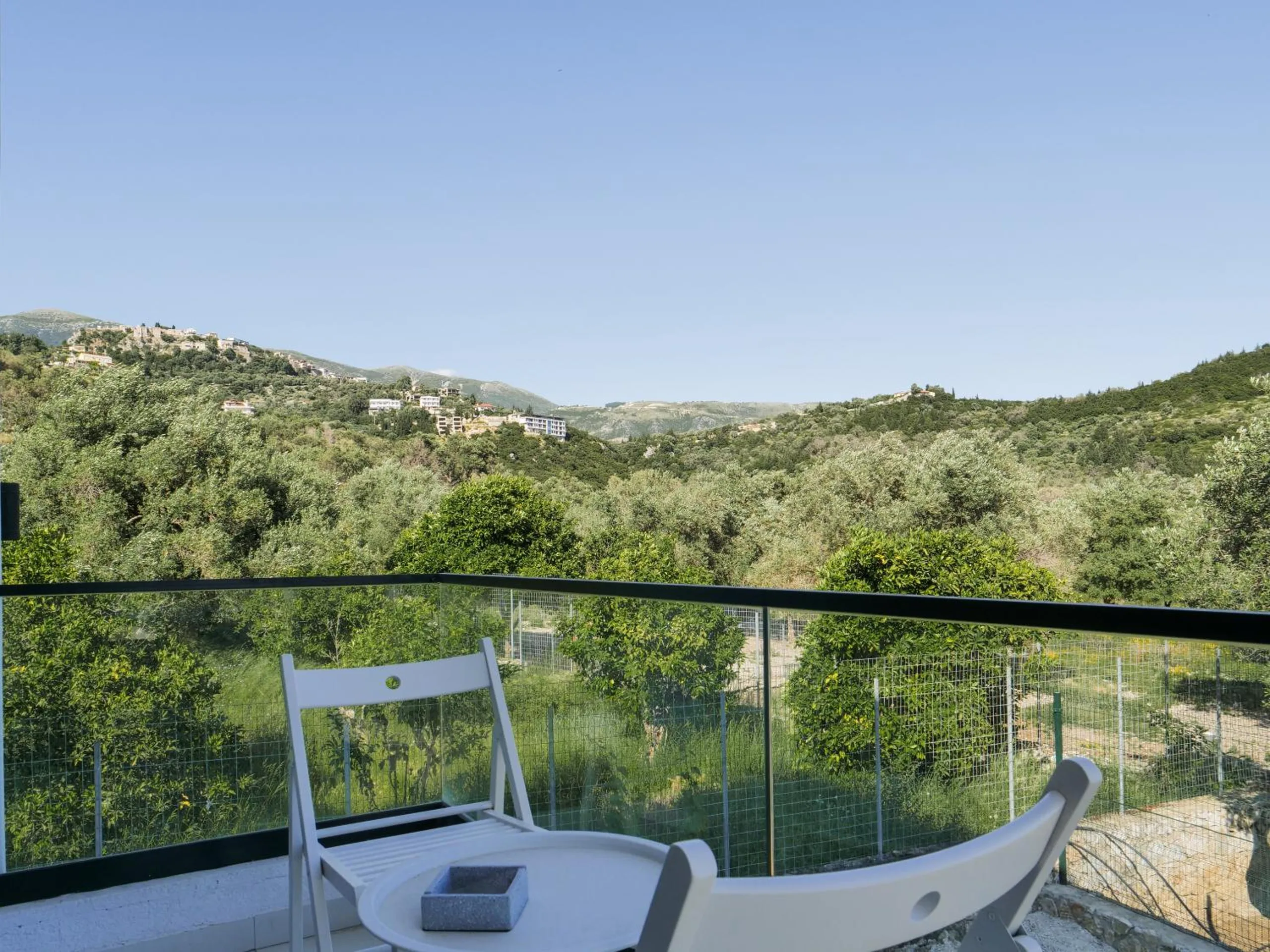 A terrace with two white chairs behind a glass rail, looking over orange and olive trees to the castle hill
