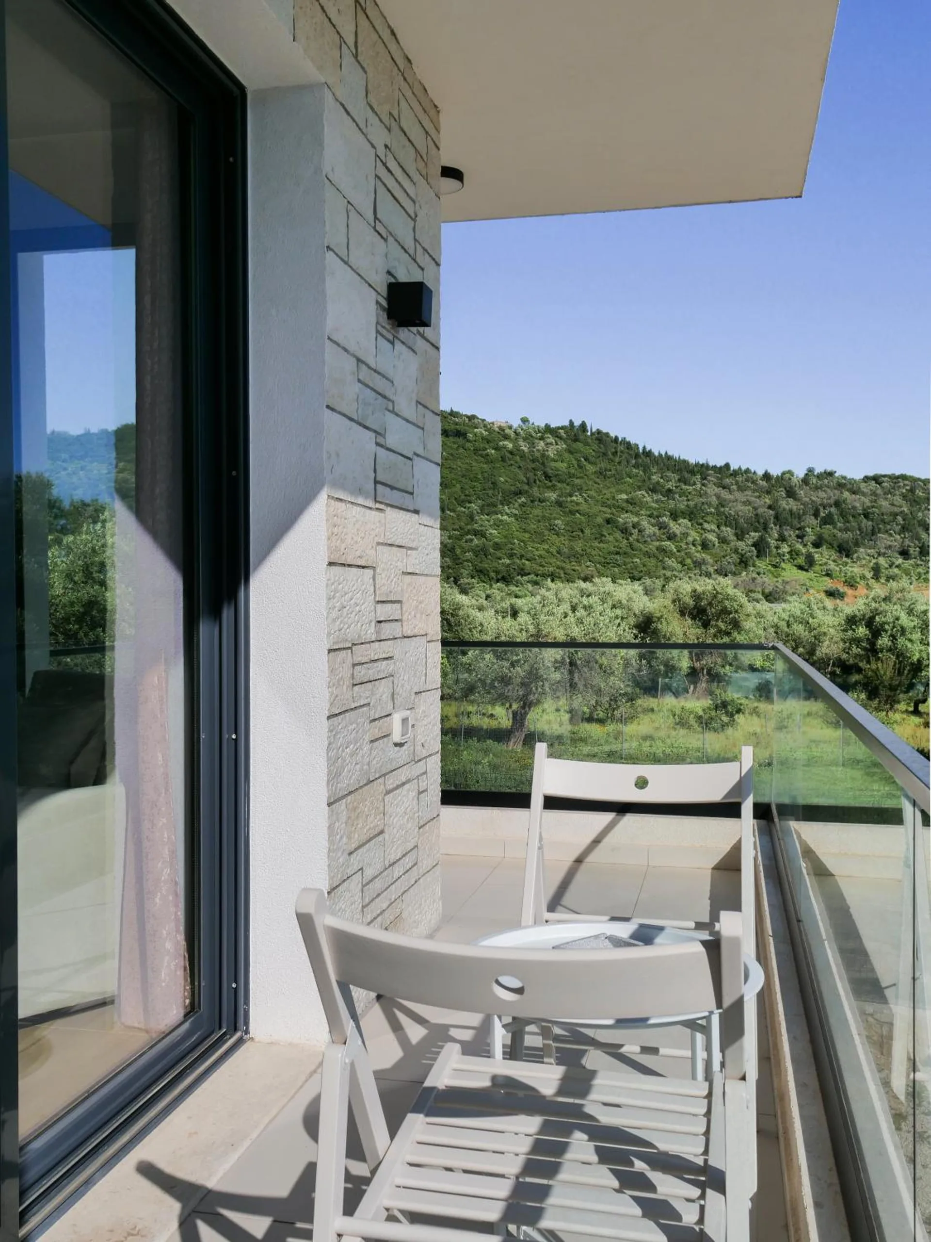 A narrow terrace with folding chairs and a glass rail beside the stone-clad wall, green hills beyond