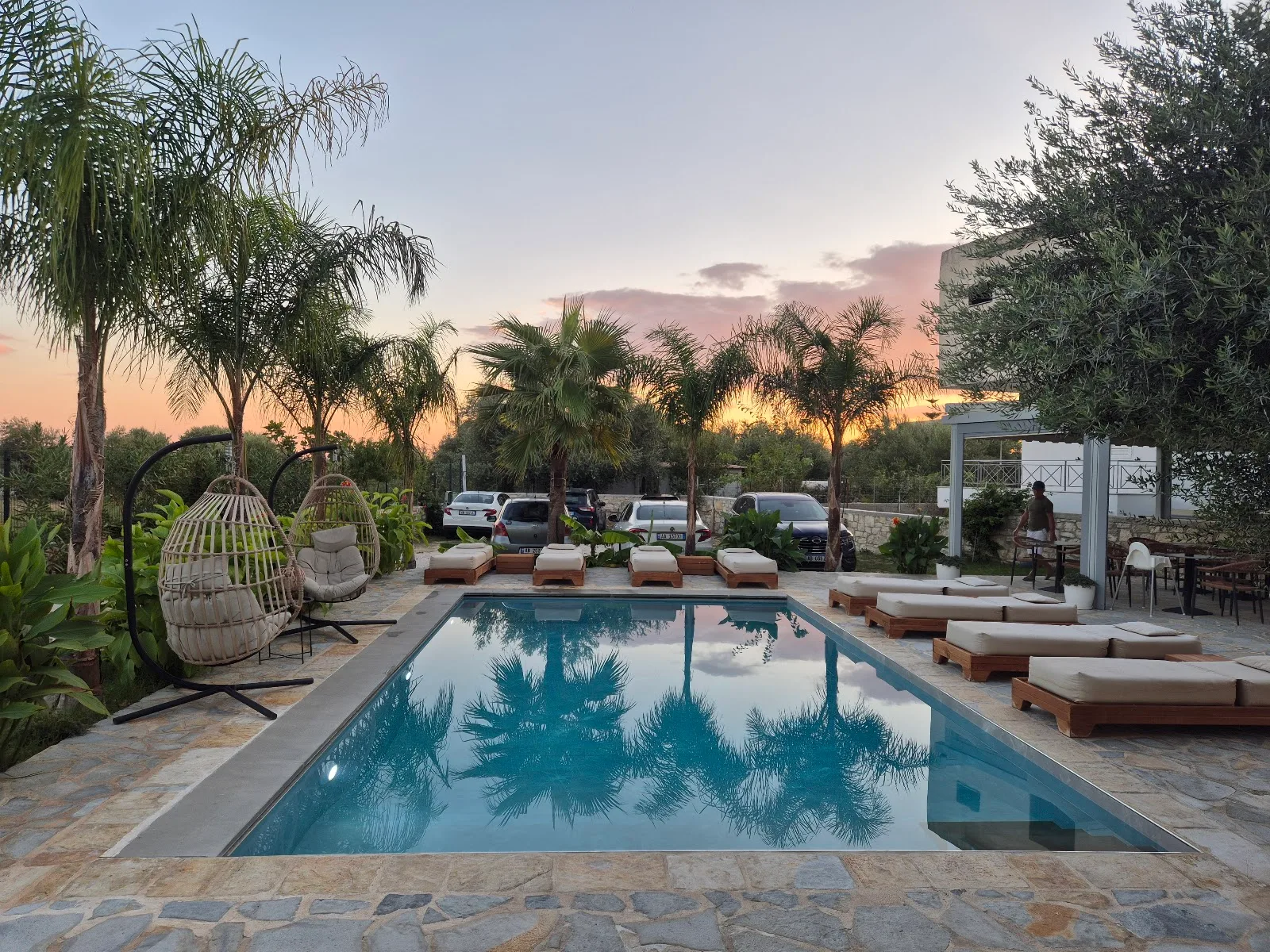 The pool at dusk under a pink sky, palms reflected in the water, hanging egg chairs and loungers around it
