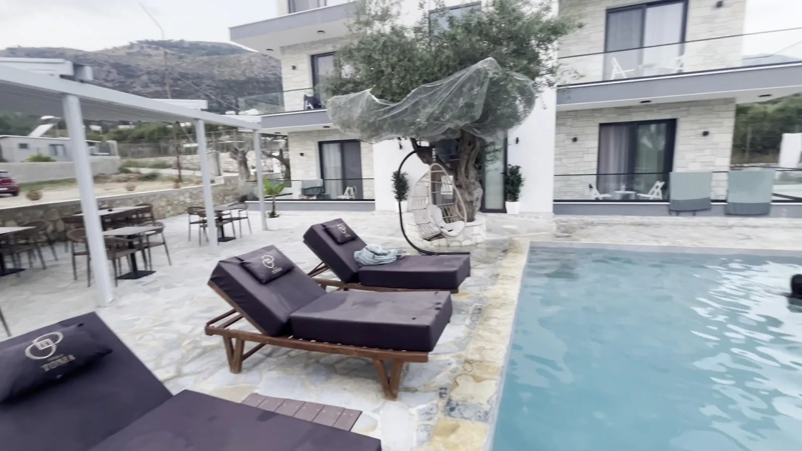 Dark loungers with gold-embroidered cushions beside the pool, an olive tree and the house behind