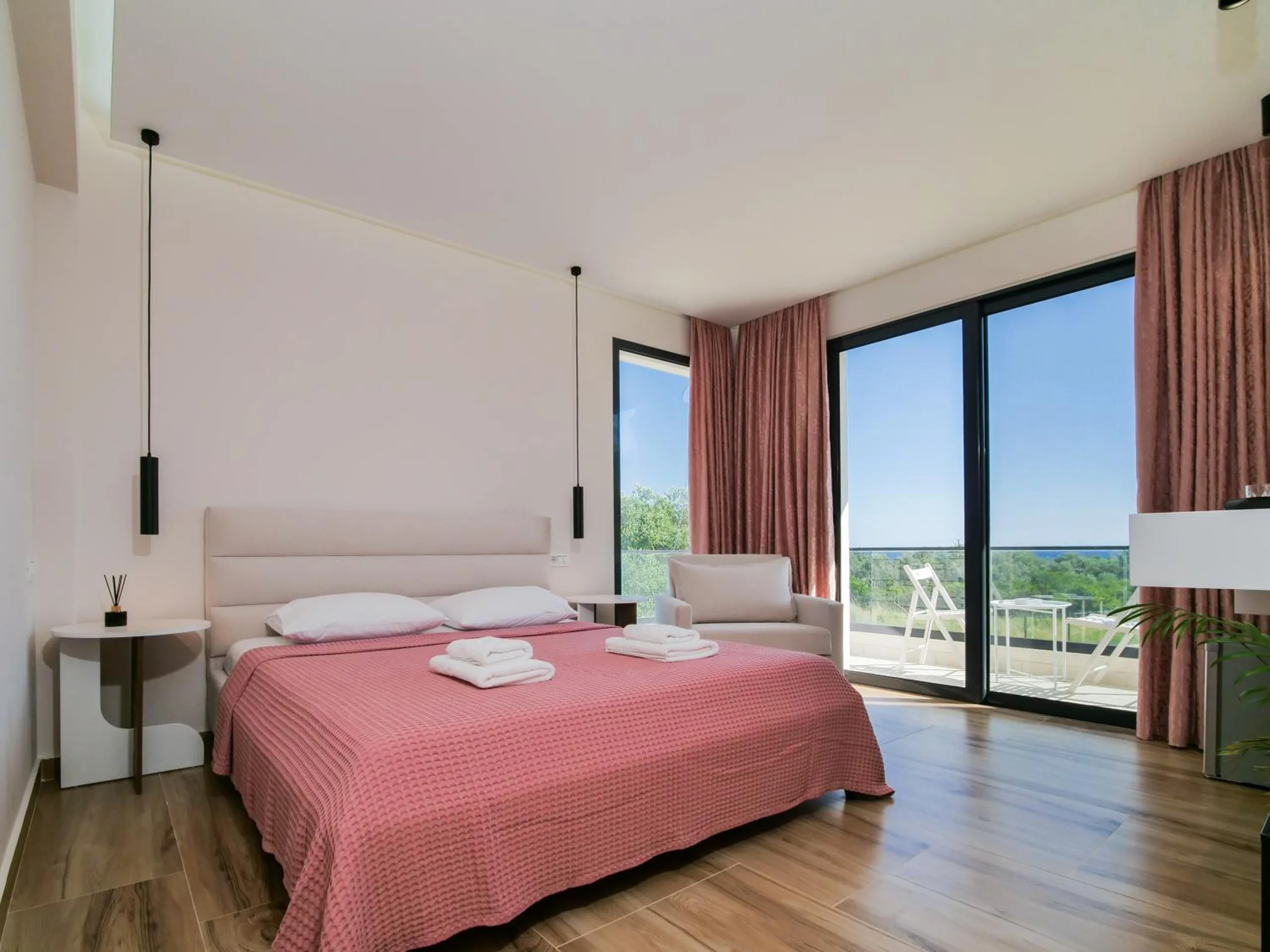 A double room with a rose bedspread, an armchair and full-height glass doors onto a terrace and greenery