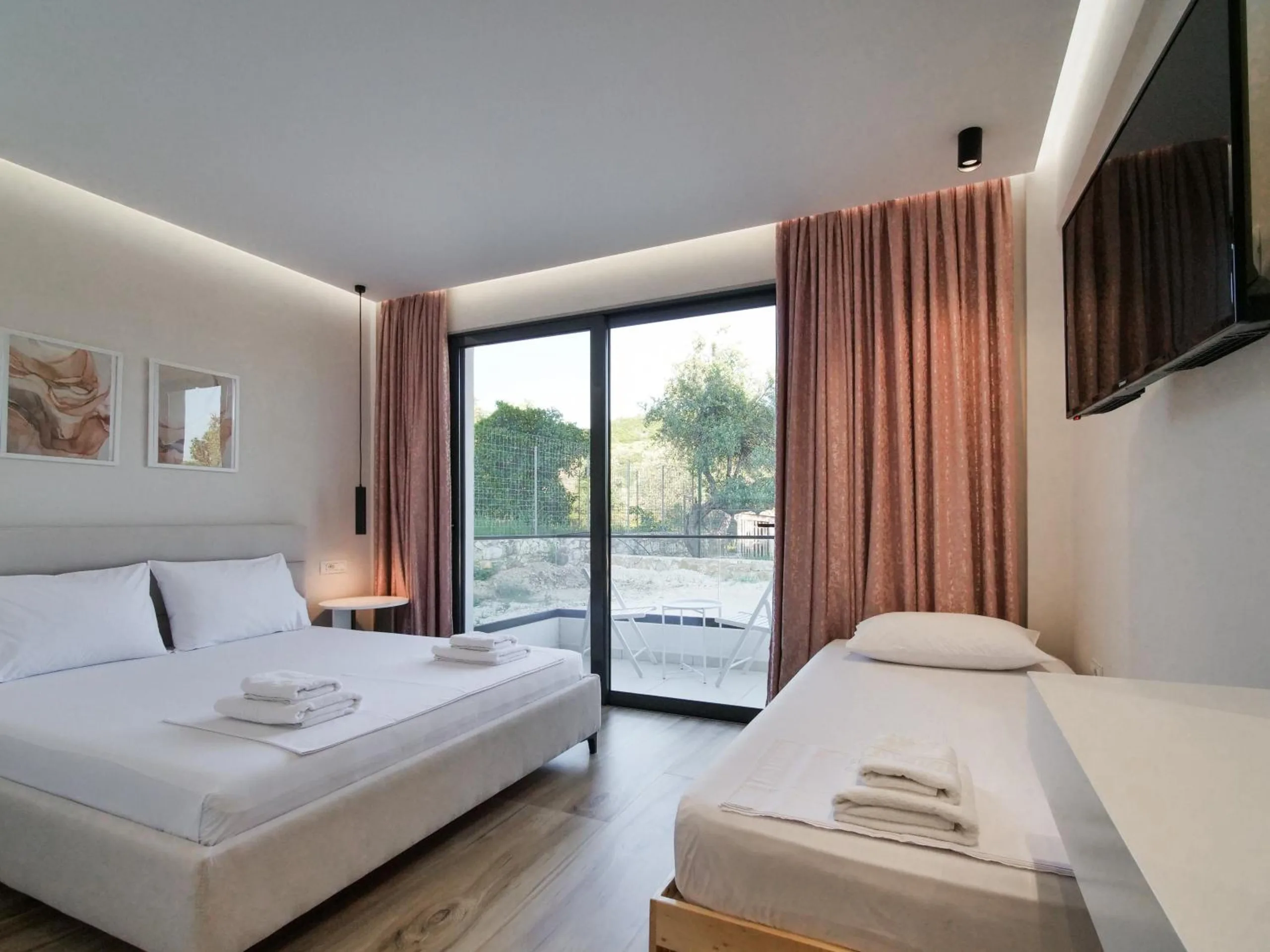 A room with a double bed and a single bed under two framed prints, rose curtains and glass doors to the olive trees