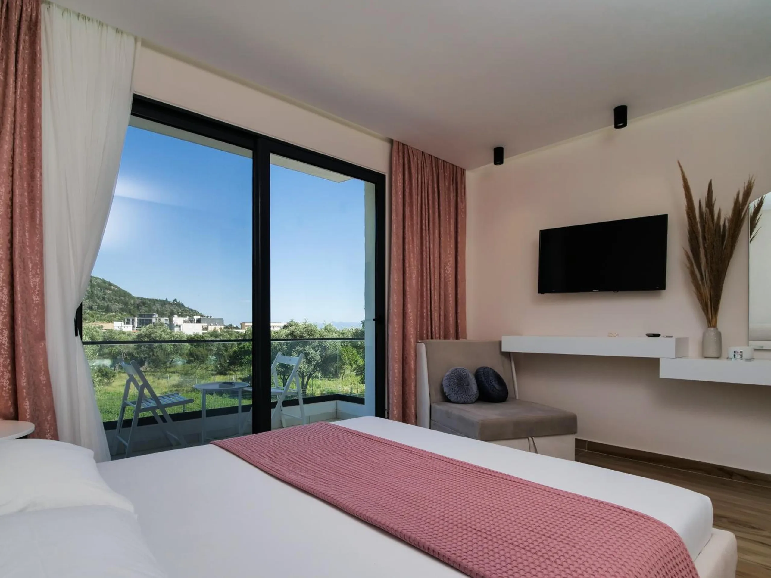 A double bed with a rose throw, glass doors open on hills and olive groves under a blue sky