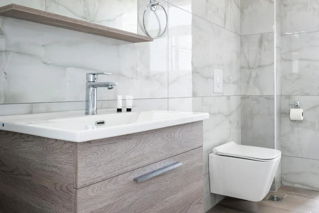 A white marble bathroom at Wait 'n Sea with an oak vanity, a wide rectangular basin, a wall-hung WC and a glass shower screen