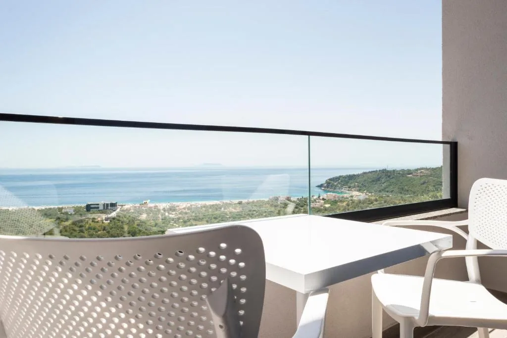A white table and two chairs on a balcony at Wait 'n Sea above the whole bay, with a low island on the horizon beyond the glass balustrade